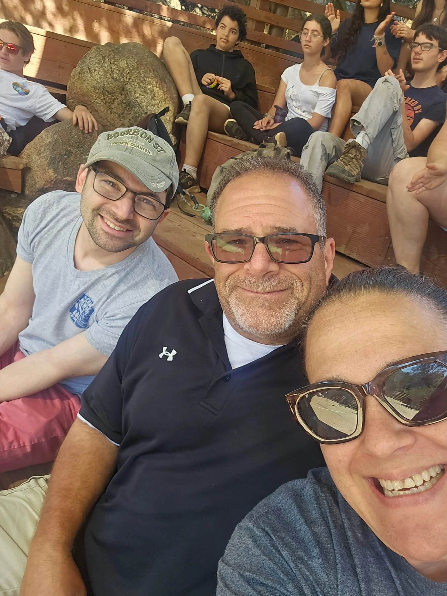A group of people sitting on wooden benches outdoors, with three individuals in the foreground taking a selfie. The person on the right, wearing sunglasses and a gray shirt, is smiling. The person in the middle, wearing glasses and a black polo shirt