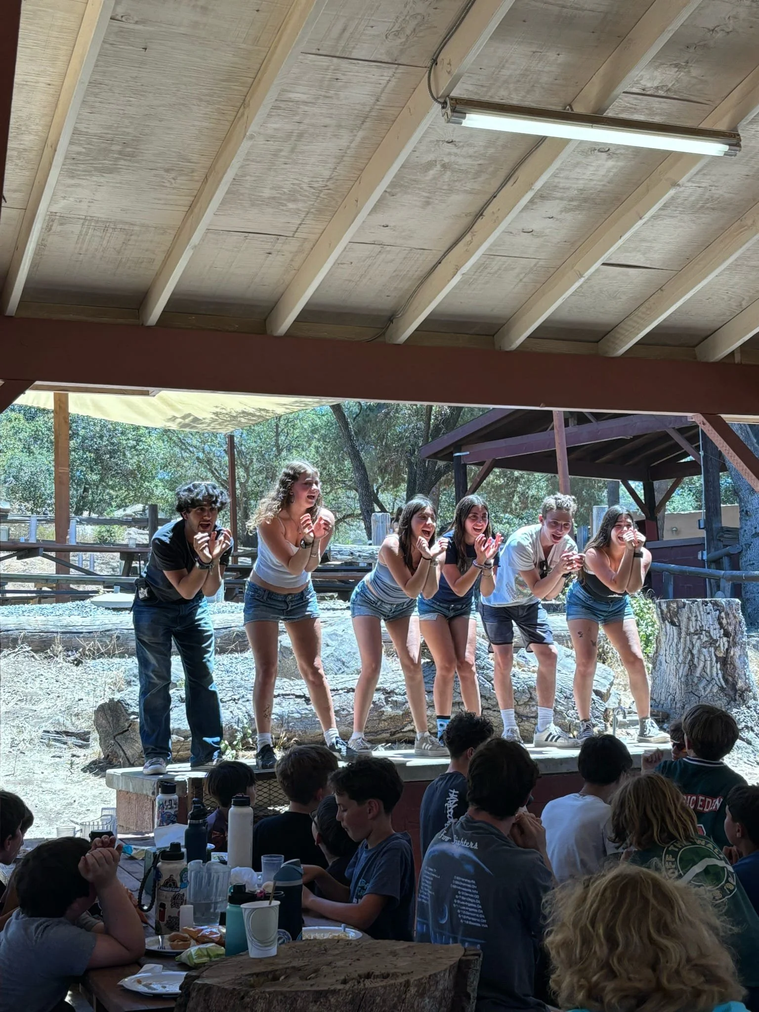 Group of children performing on a small stage at an outdoor venue, with audience of children seated at tables watching.