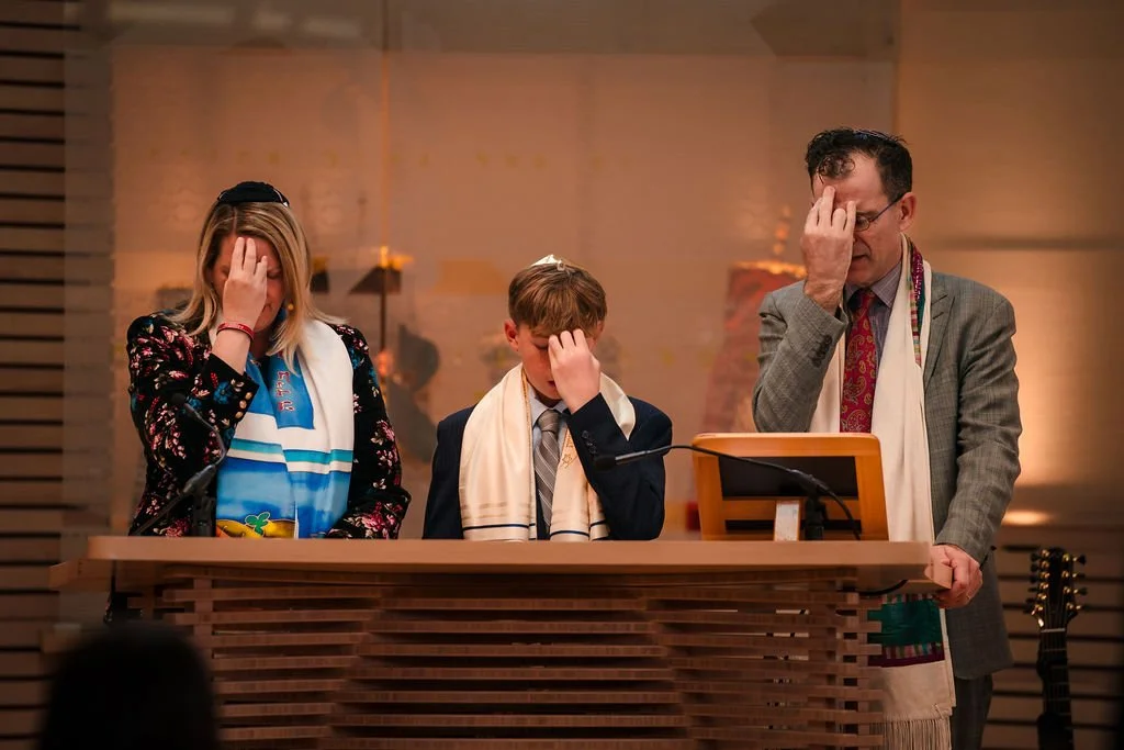 A family praying at a synagogue during a religious ceremony, each with hands covering their faces and wearing prayer shawls.