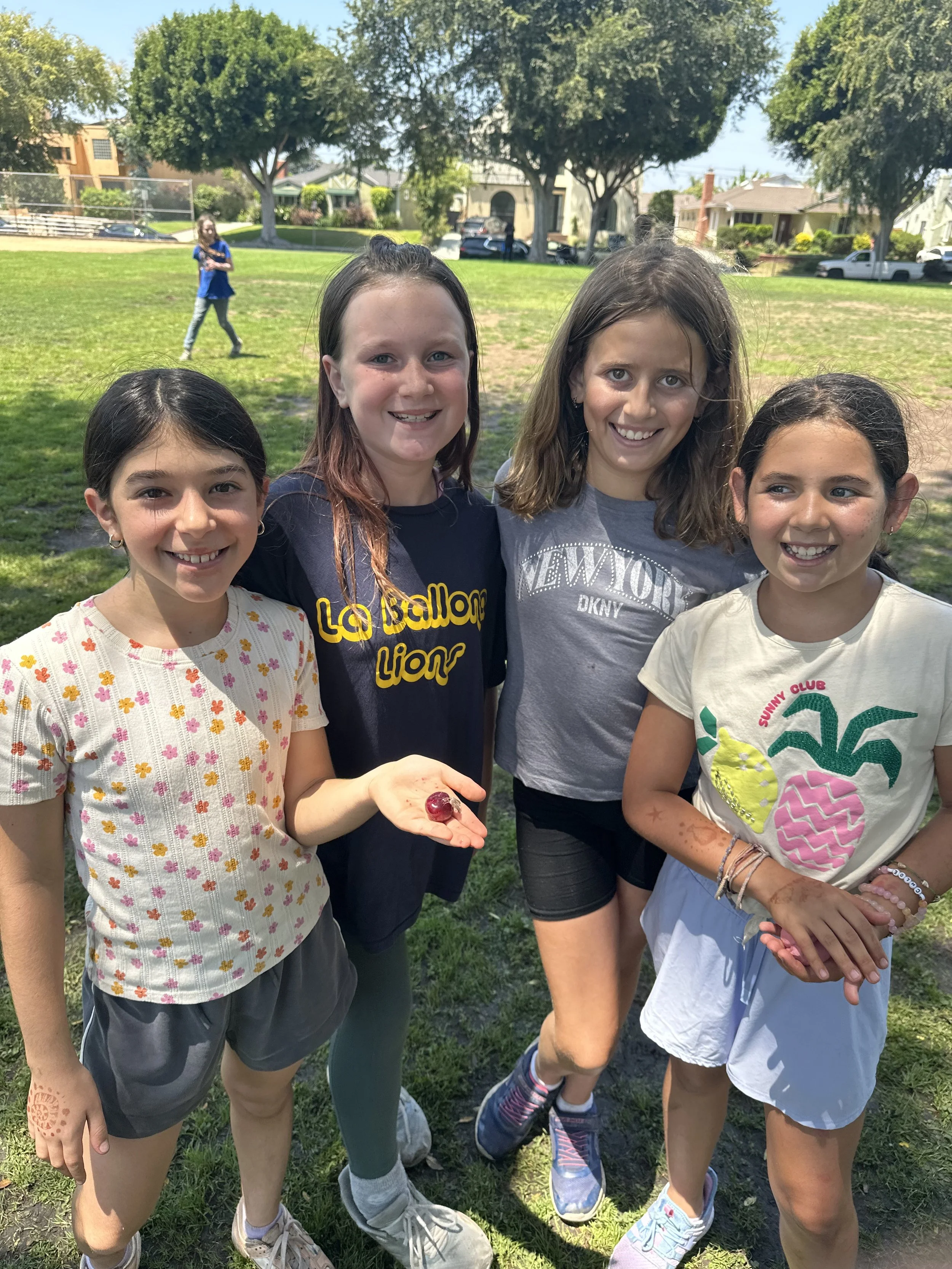 Four girls standing together in a park on a sunny day, with one girl holding a cherry in her hand. Other children are playing in the background.