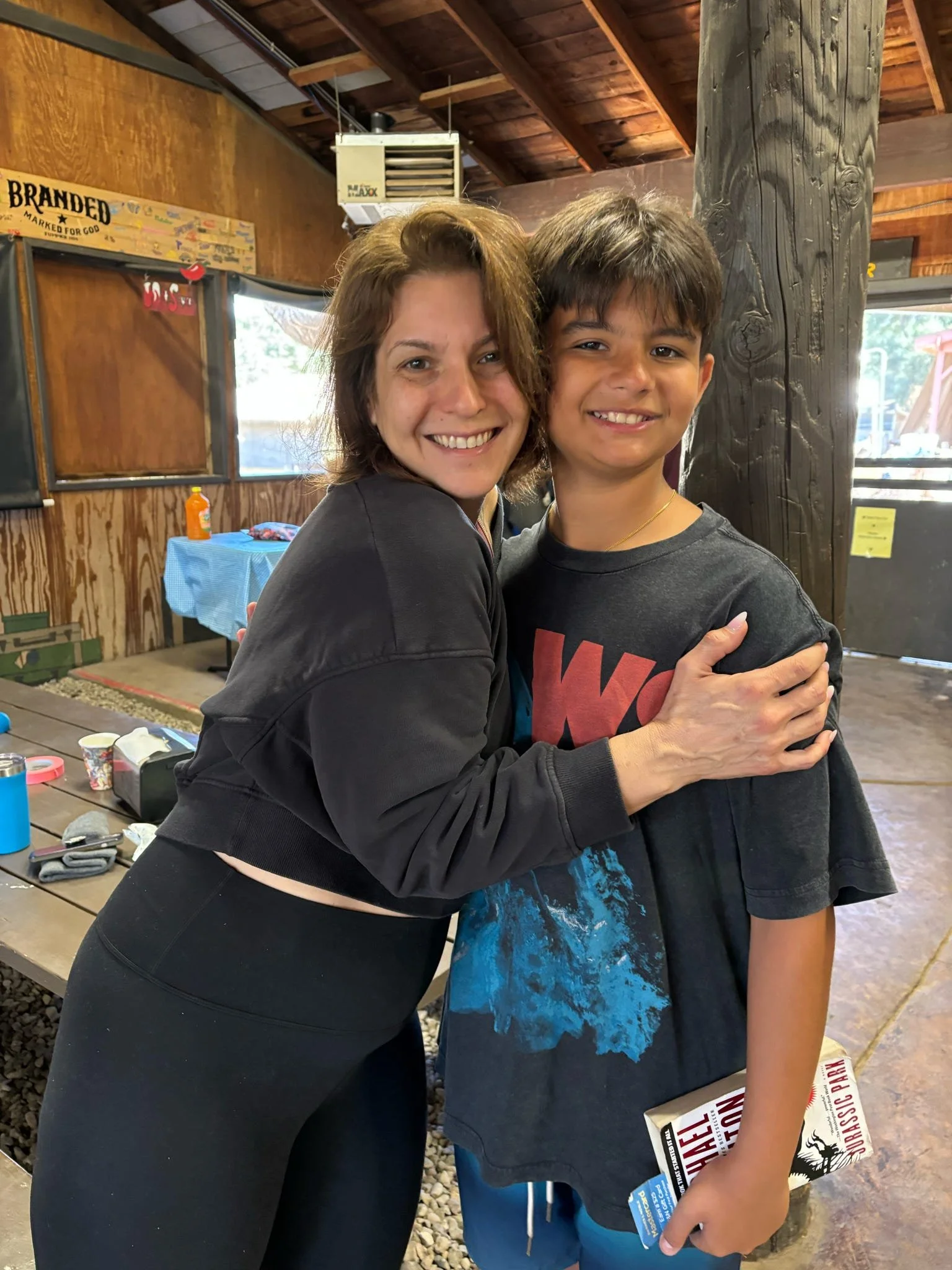 A woman and a young boy hugging and smiling inside a wooden building, with a table and various items in the background.