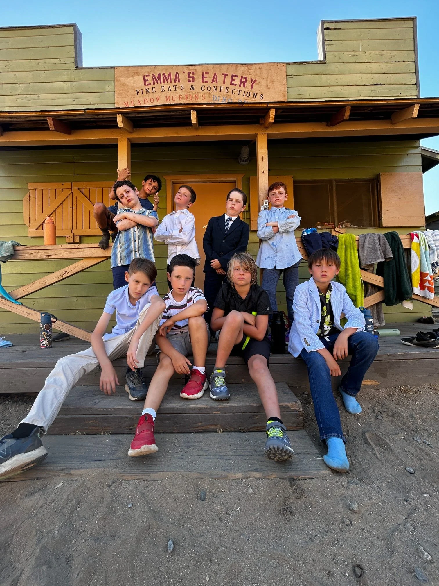 Group of children sitting and standing on the front porch of a wooden house, some with crossed arms, others posing with a rebellious attitude, with laundry hanging on the side and a sign that reads 'Emma's Eatery.'