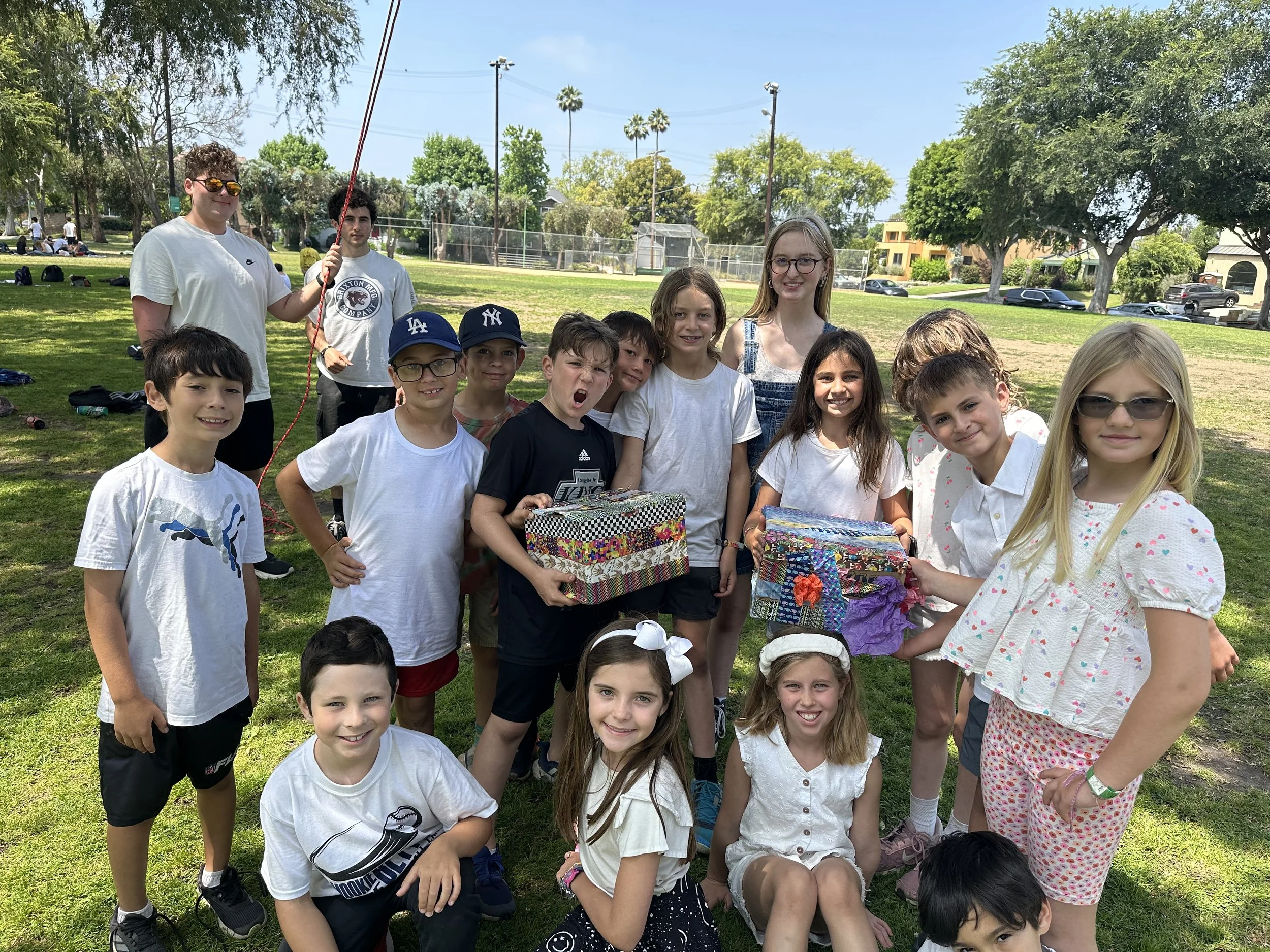 Group of children and one adult in a park, with some holding decorated boxes and balloons, sunny day with trees and park equipment in the background.