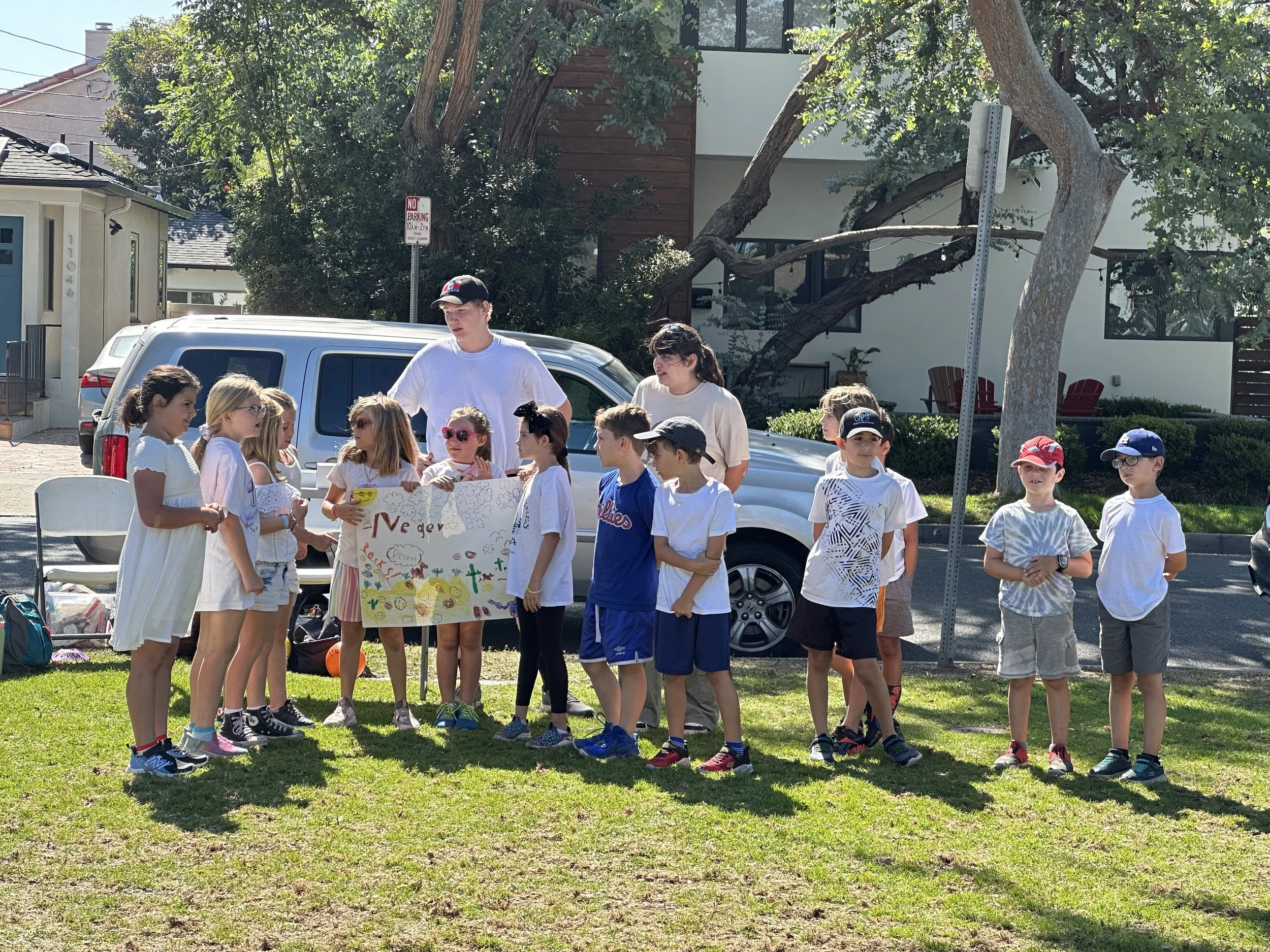 A group of children and two adults standing on grass outdoors, holding a colorful sign with drawings and words, with parked cars and trees in the background.