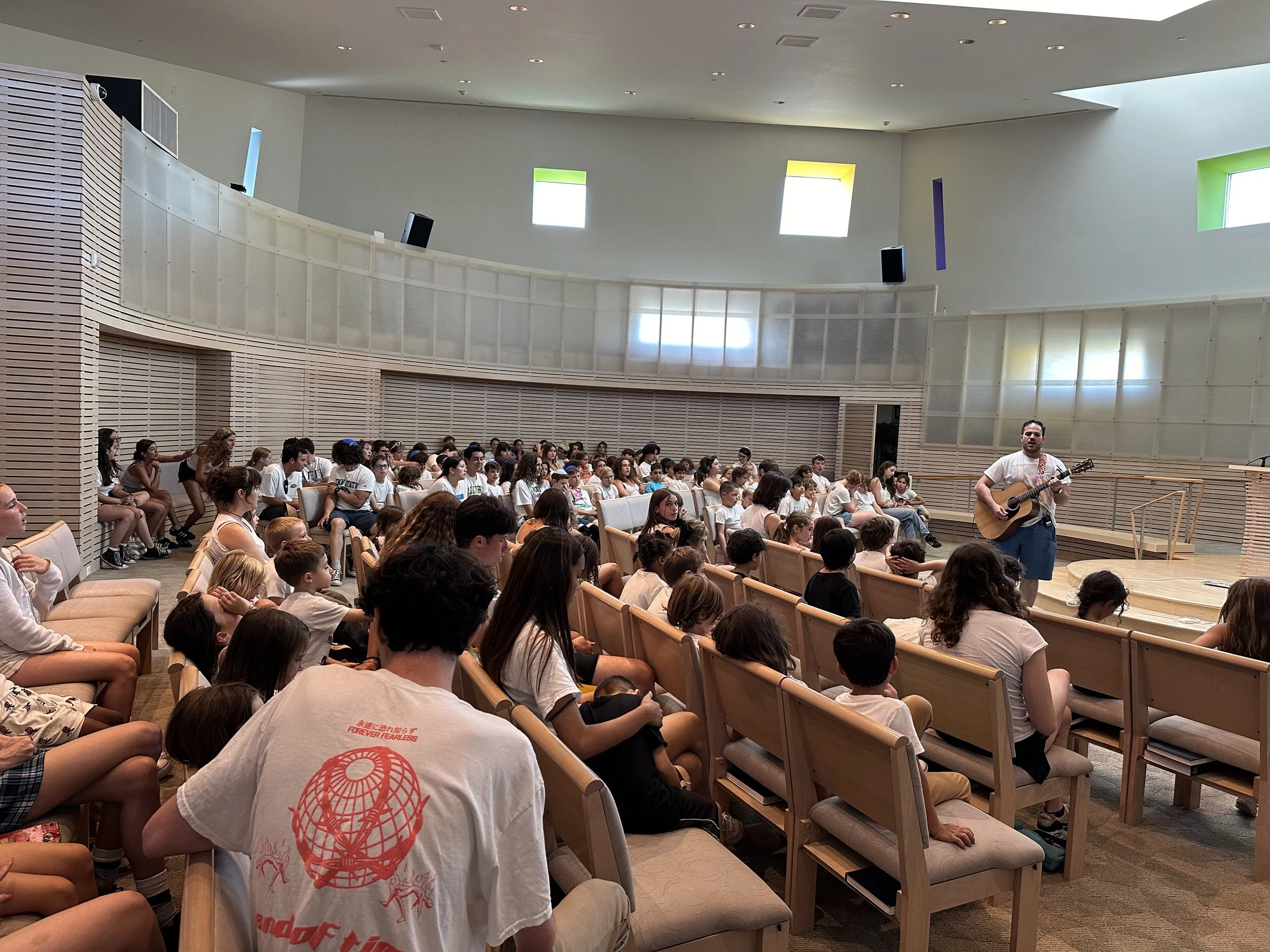 A group of children and teenagers sitting in an auditorium, listening to a man playing guitar and singing on stage. The auditorium has modern design with wooden accents and colored window panels.