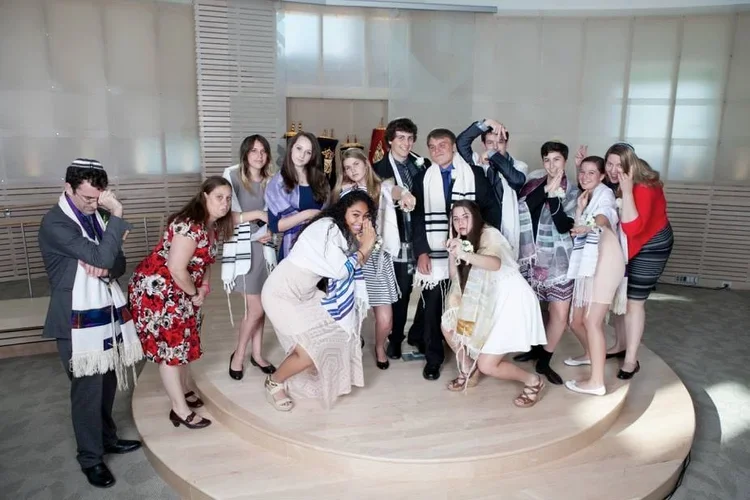 Group of young people dressed in winter clothing, gathered on a stage in a room with large windows, celebrating Hanukkah with menorahs in the background.