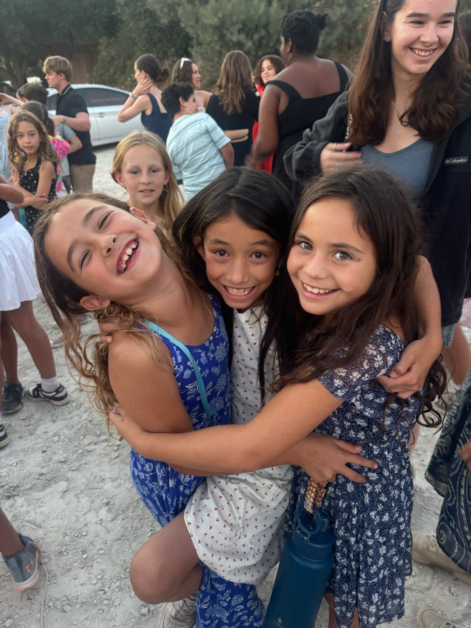 Three young girls hugging and smiling at an outdoor gathering with other children and adults in the background.