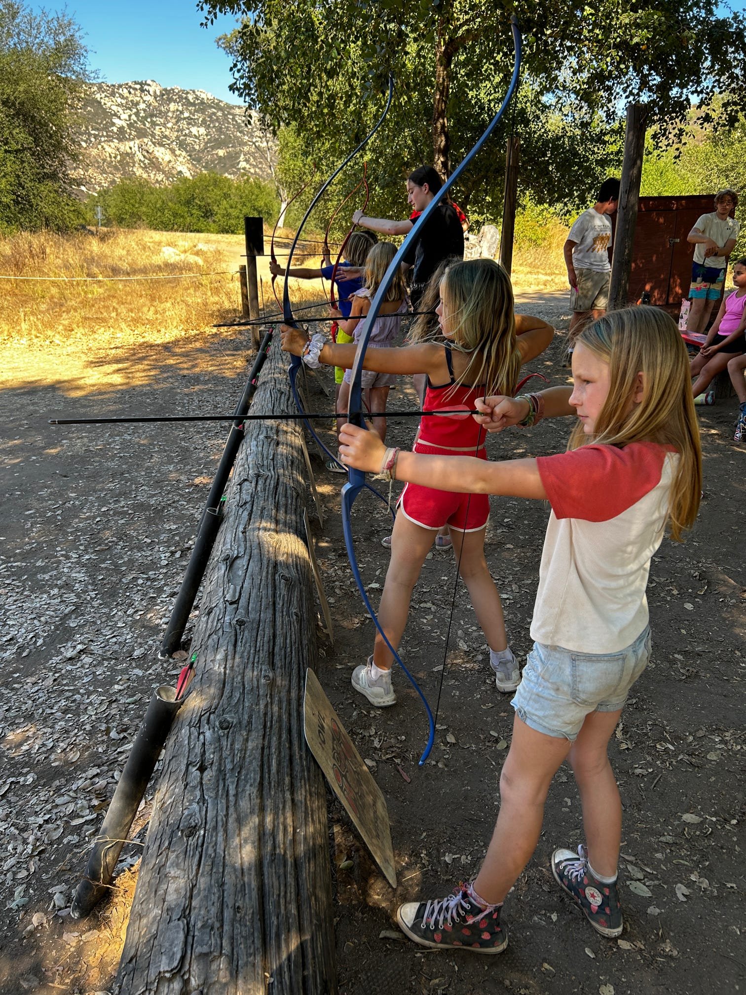 Children participating in an outdoor archery activity, pulling bows and aiming arrows.