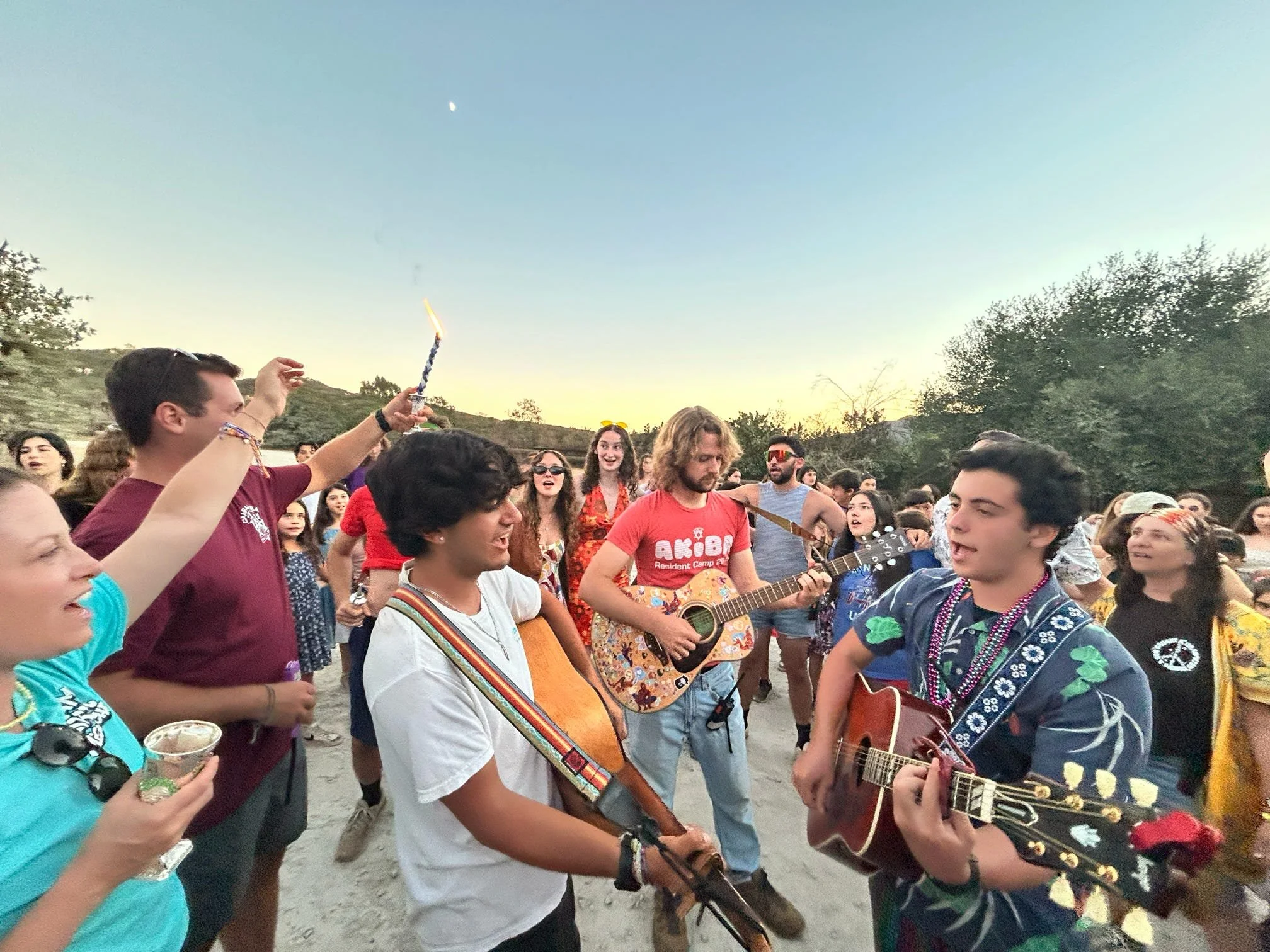 Group of young people gathering outdoors at sunset, some playing guitars, singing, and dancing, with festive and casual attire.