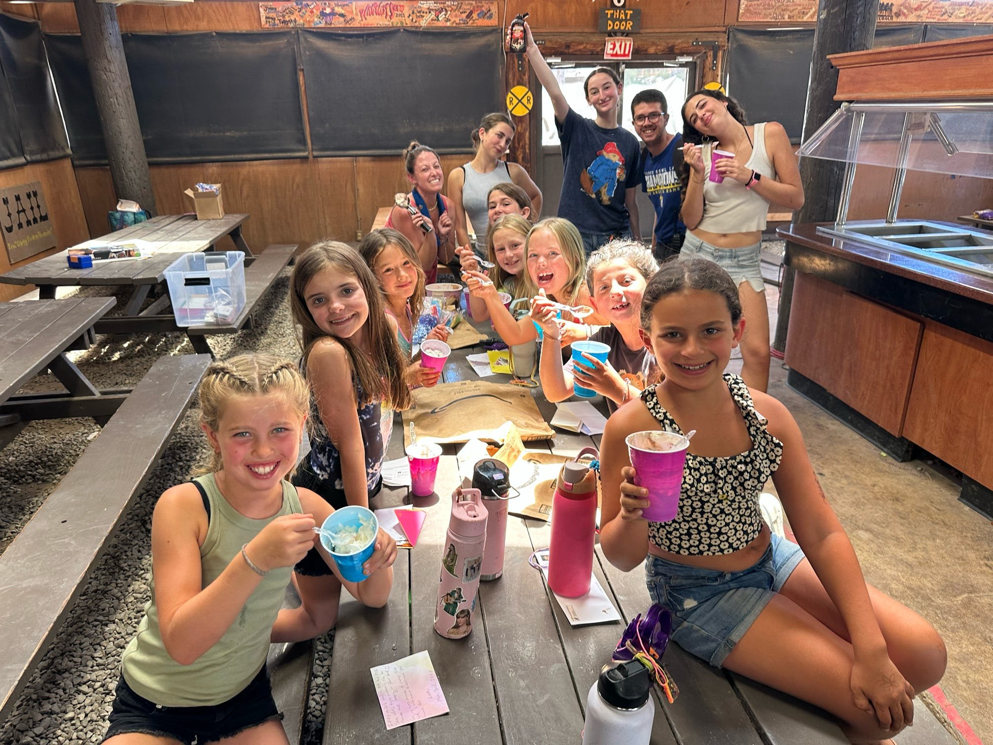 Group of children and teenagers enjoying ice cream and drinks at a picnic table inside a rustic building, with some of the older individuals standing in the background.