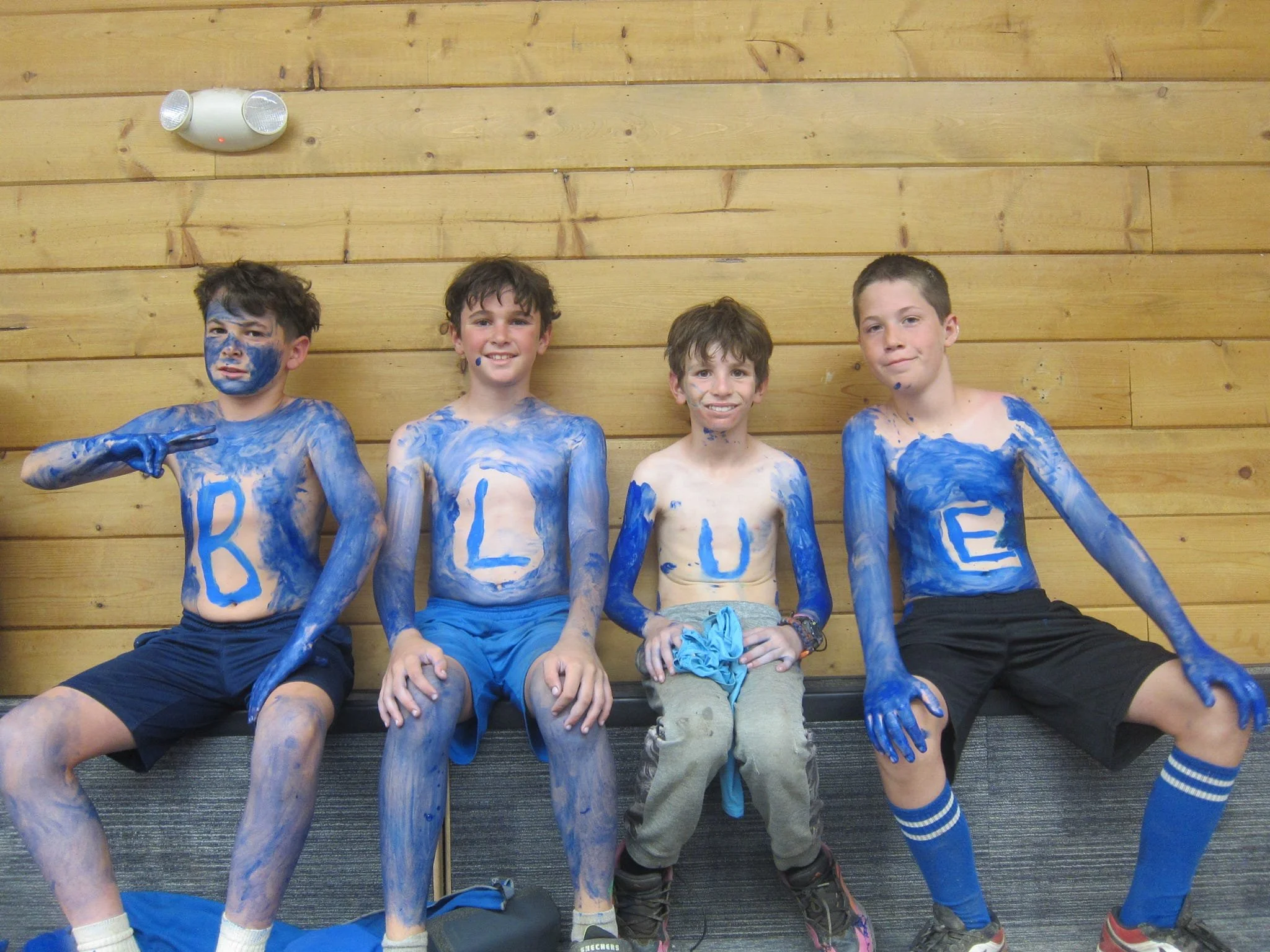 Four boys sitting against a wooden wall, painted with blue body paint spelling 'BLUE'. They are smiling, and their bodies and faces are painted in blue.