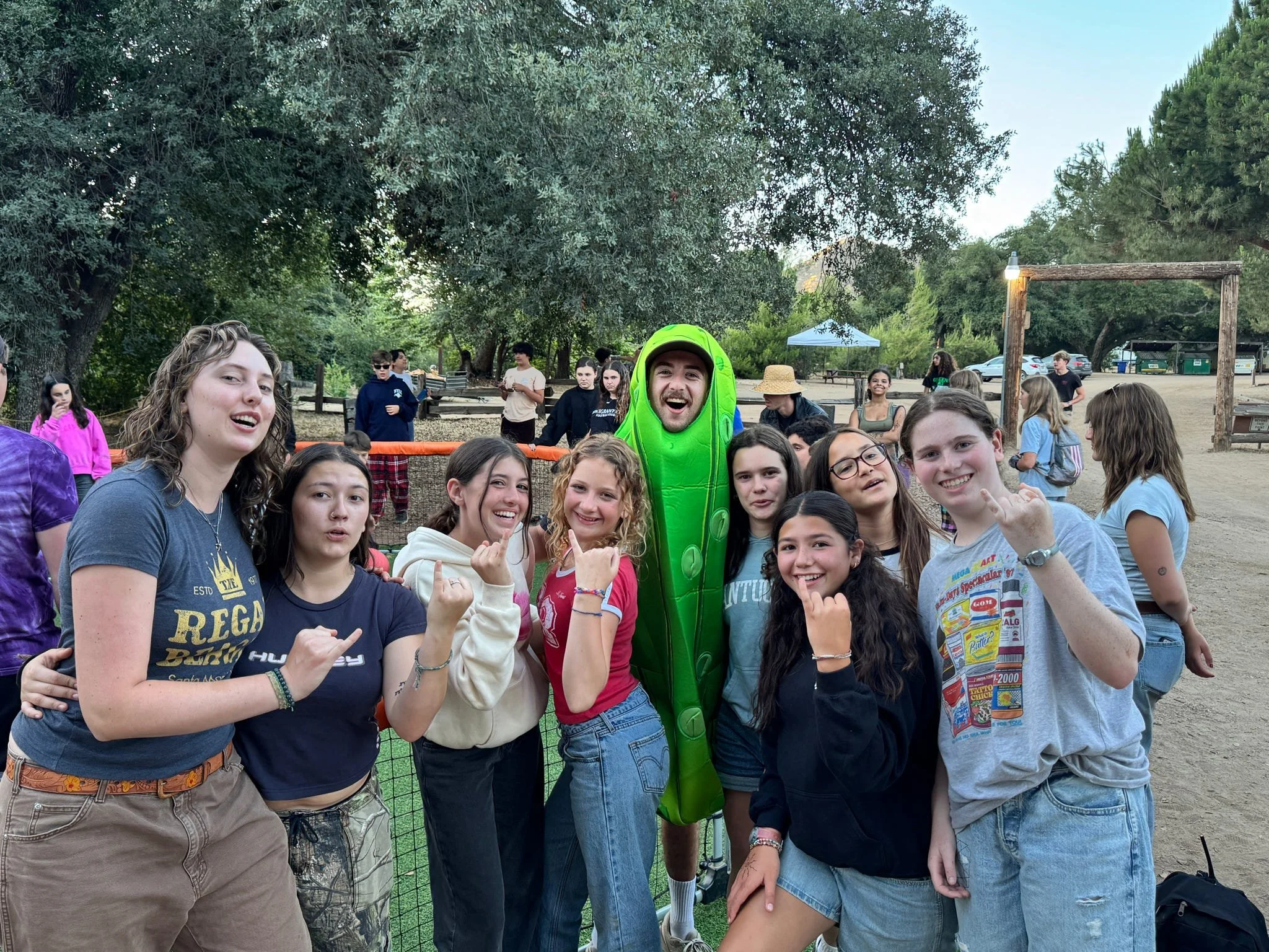 Group of diverse children and teenagers posing with a person in a pickle costume outdoors in a park.