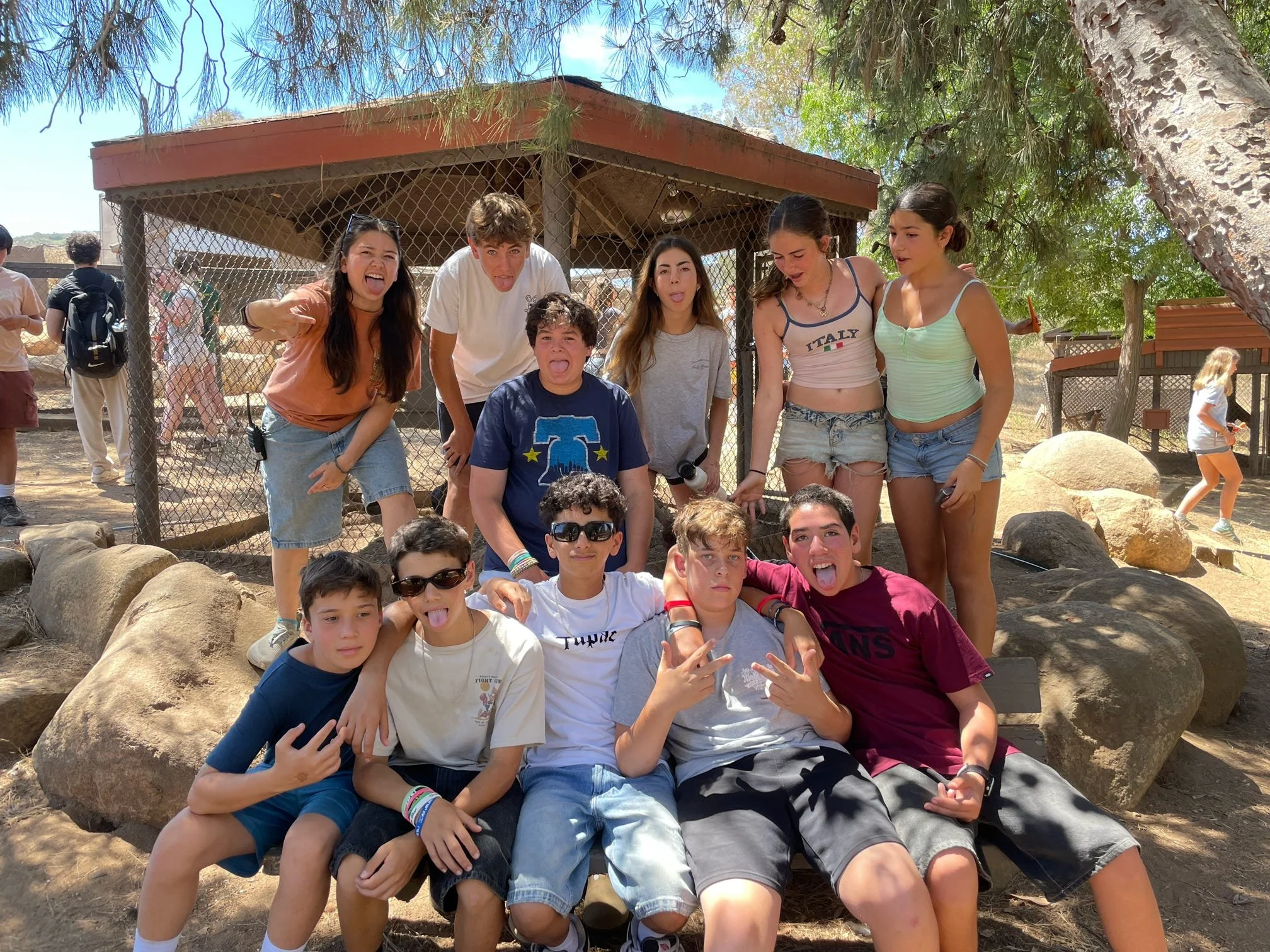 Group of teenagers posing outdoors with rocks and trees, some making silly faces, under sunny weather.