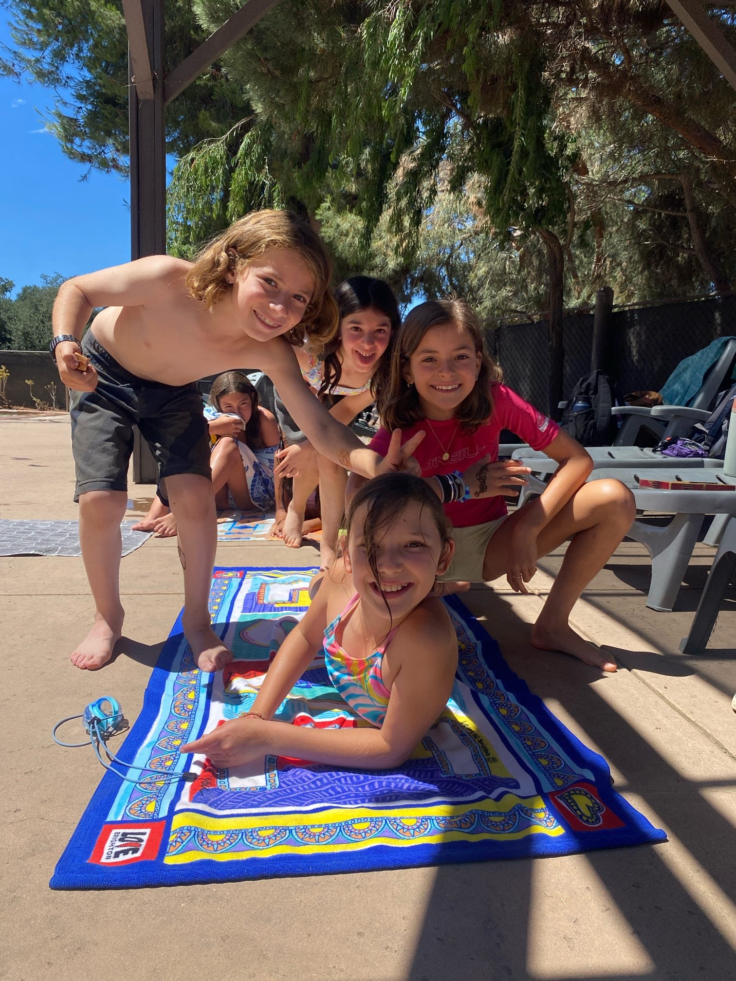 Group of children playing and posing on a colorful towel outdoors, smiling and making peace signs.