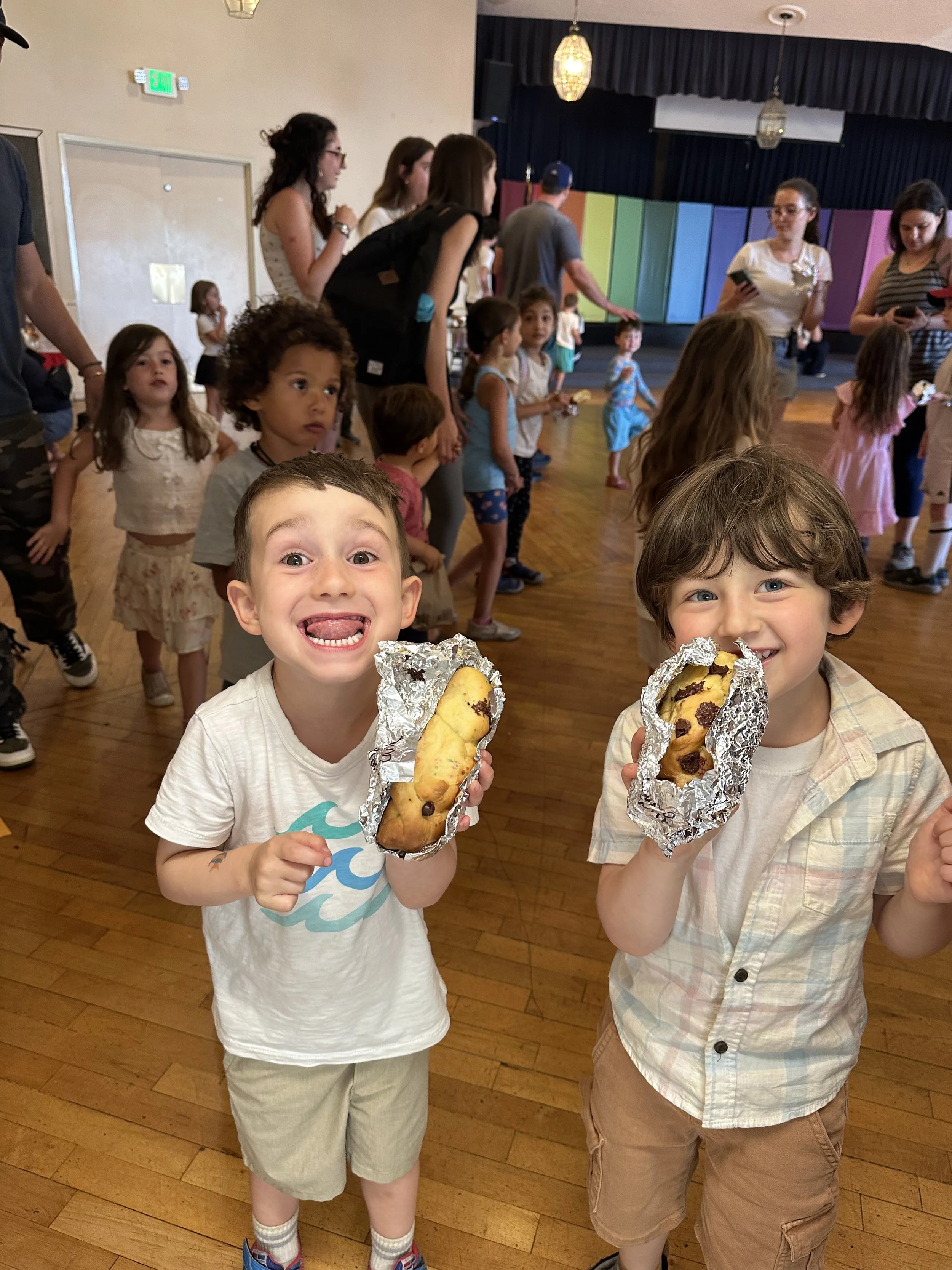 Two young boys smiling and holding large chocolate chip cookies wrapped in aluminum foil, indoors at a children's event with other kids and adults in the background.