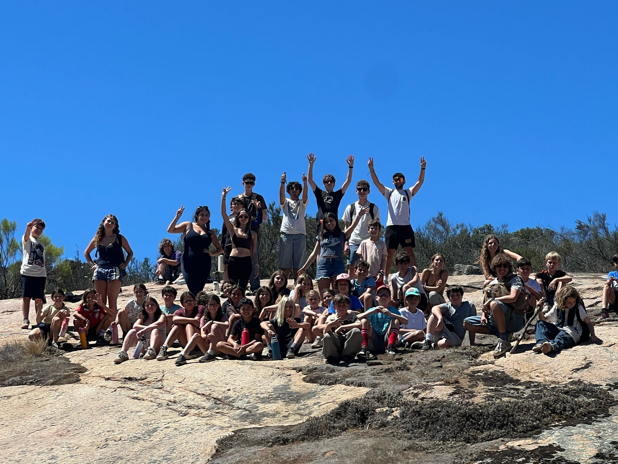 Group of children and teens on a rocky outdoor terrain under a clear blue sky, some standing and some sitting, posing for a photo.