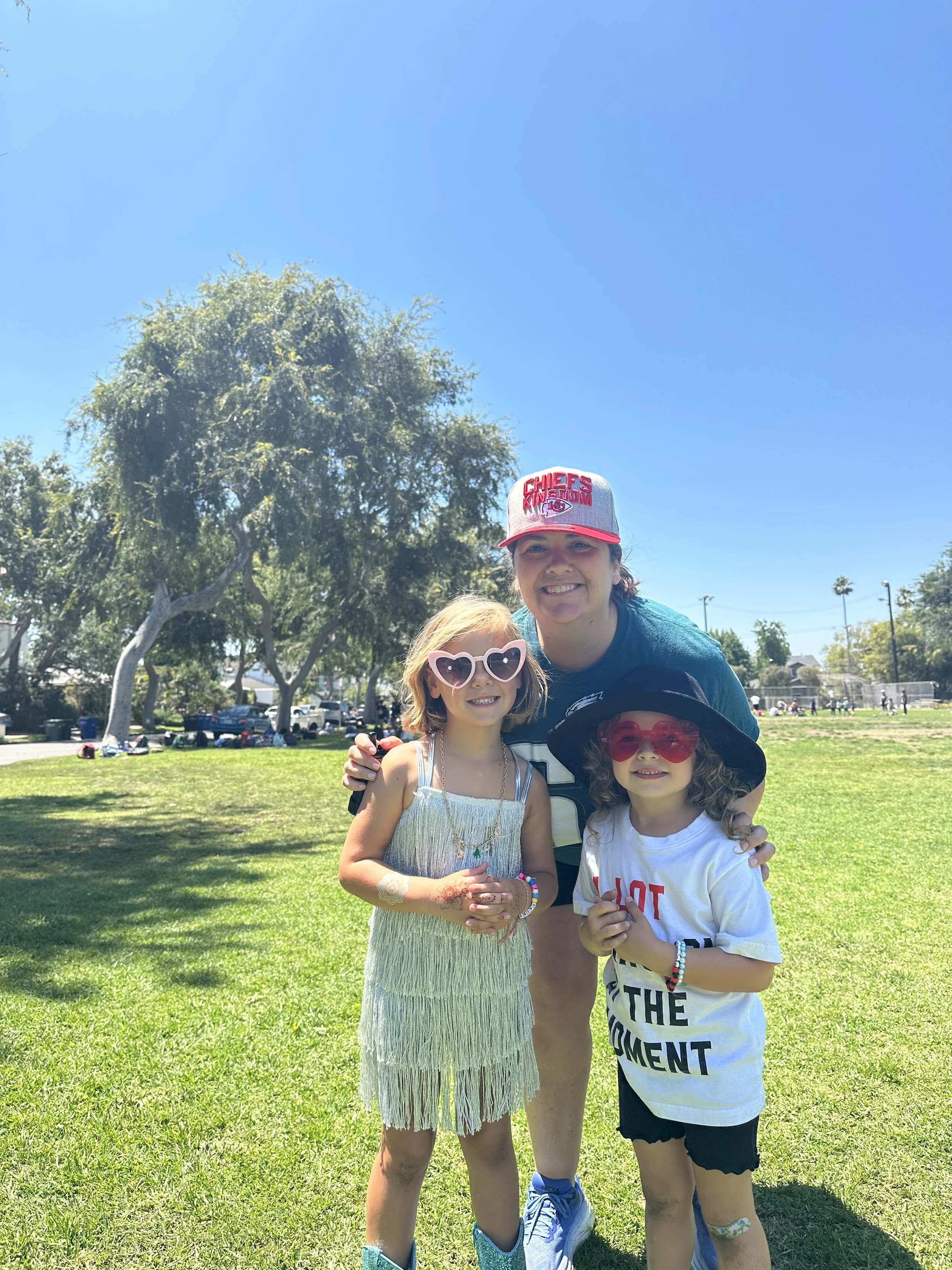 A woman and two young girls are outdoors on a sunny day, smiling and posing for a photo in a park with green grass and trees in the background. The woman is wearing a cap, a blue shirt, and has her arms around the girls. The girl on the left has blon