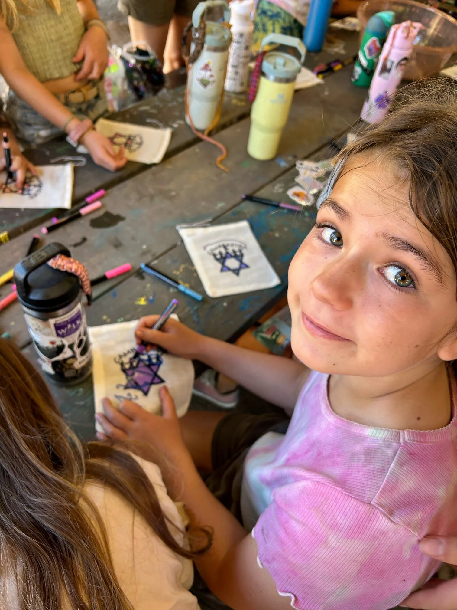A young girl with curly hair and a pink shirt smiling while coloring a tote bag with a purple hat and star design at a craft table, surrounded by markers, water bottles, and other kids' art projects.