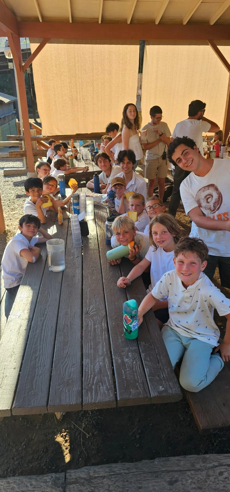 A group of children and teenagers sitting and standing around a wooden picnic table outdoors, smiling and holding water bottles and snacks, under a shaded canopy with a background of a wooden structure.