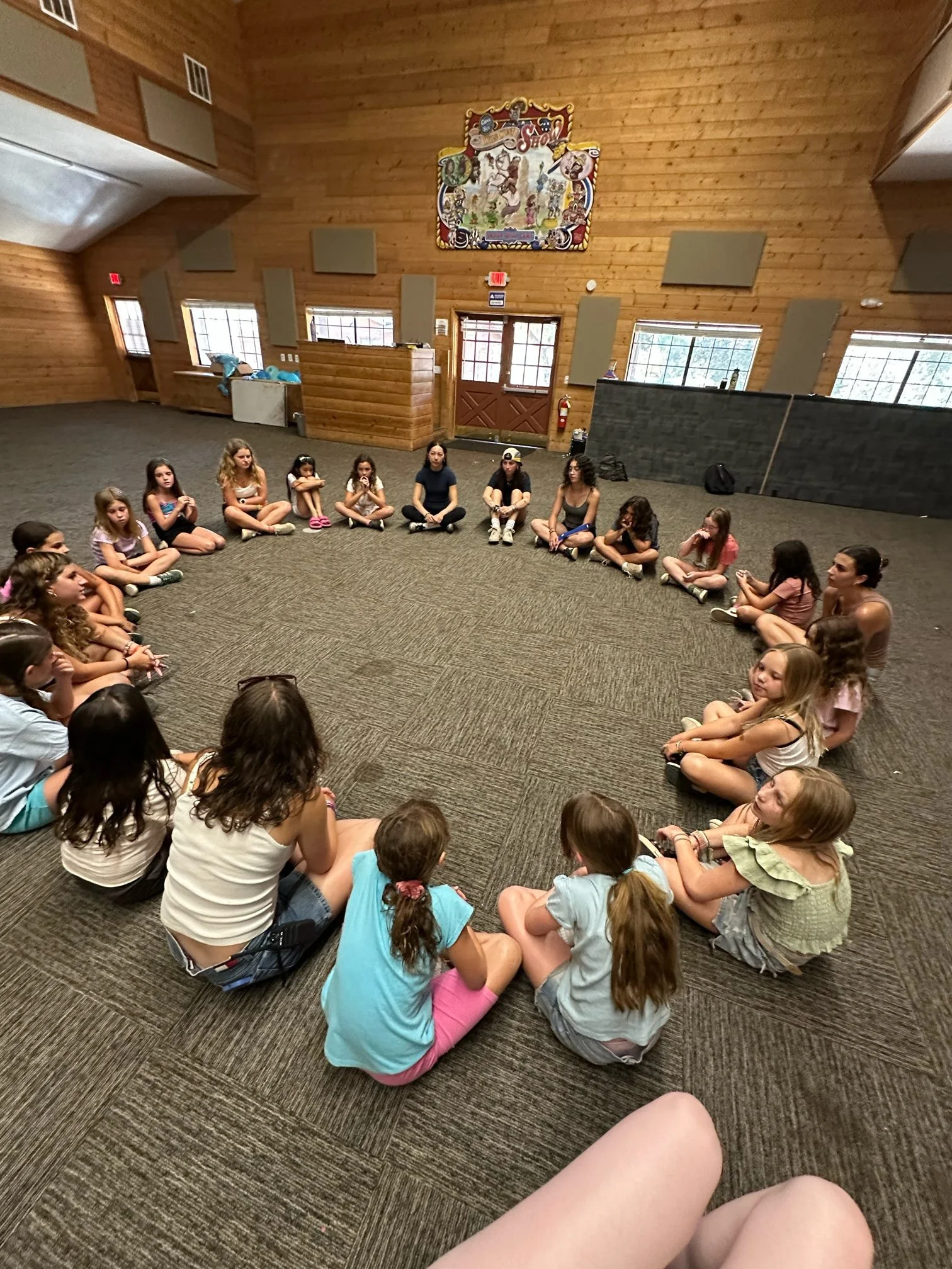 Group of children sitting in a circle in a large wooden-floored room with windows, walls, and colorful decorations.