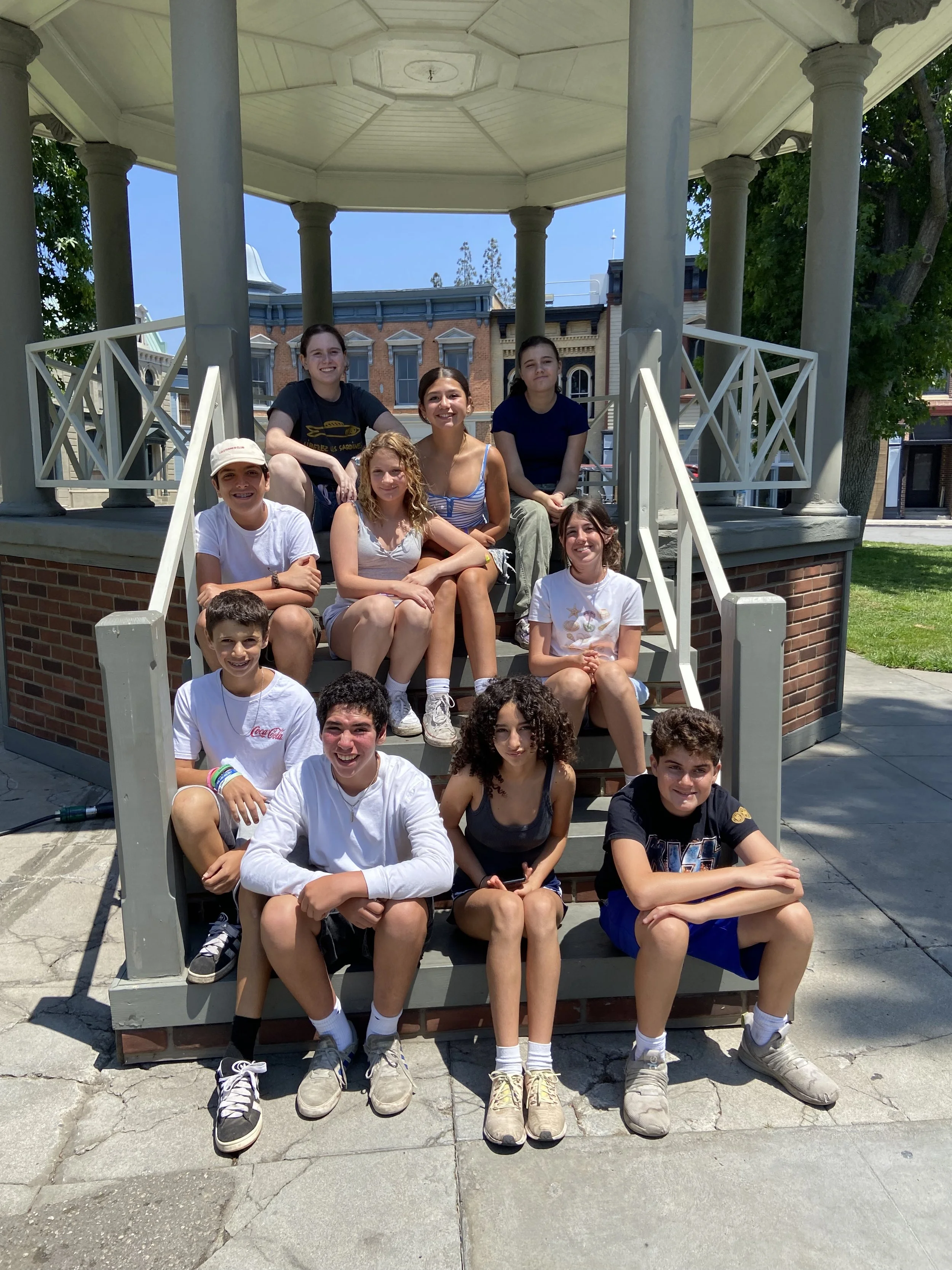 A group of thirteen kids sitting on the steps and under the roof of a gazebo in a park on a sunny day.