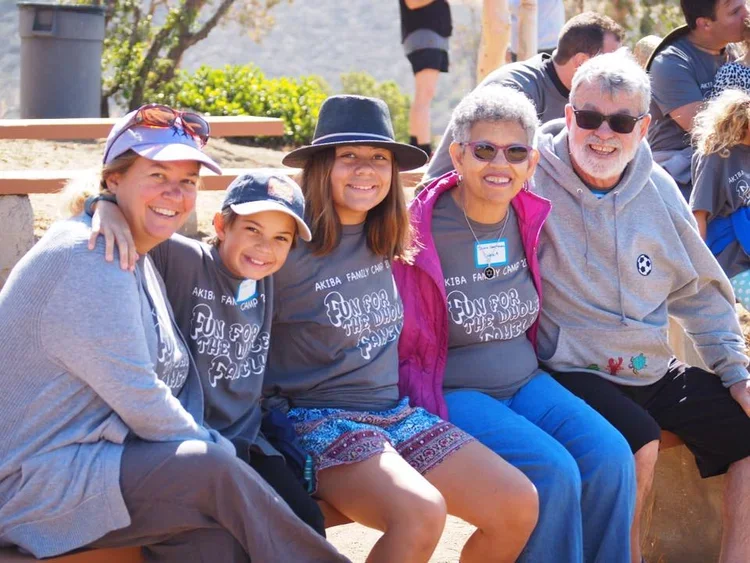 A group of five smiling people, including children and adults, sitting outdoors on a bench during a sunny day, wearing casual clothes and sunglasses.