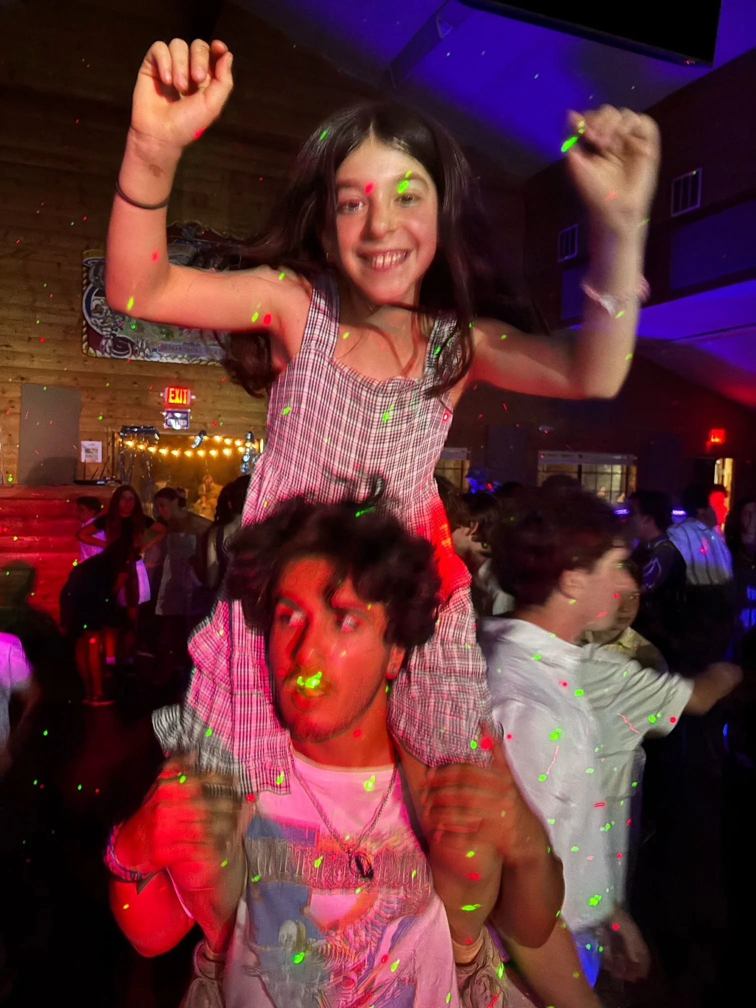 A young woman is sitting on a man's shoulders at a lively party or dance event with colorful lights. She is smiling and has her arms raised. The man, with curly hair, appears to be dancing with his fists clenched. Many people are in the background en