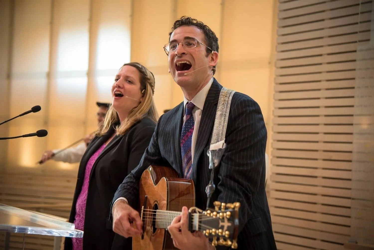 Man in a suit playing guitar and singing at a panel discussion or conference, with woman beside him also singing.