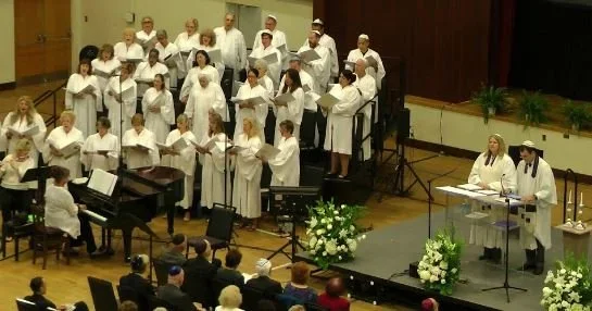 Choir group performing with a conductor and pianist on stage during a concert.