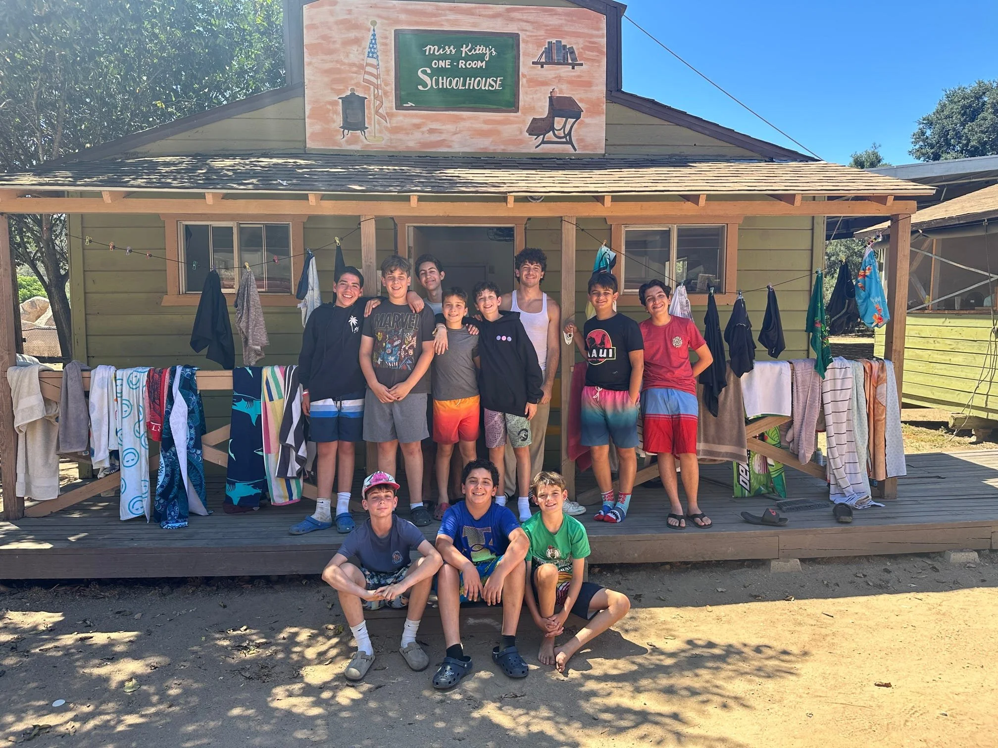 A group of boys is standing and sitting in front of a small green one-room schoolhouse with laundry hanging on a line outside. The schoolhouse has a sign that reads 'Miss Kitty's One-Room Schoolhouse' with an American flag, a bell, and chalkboard ill
