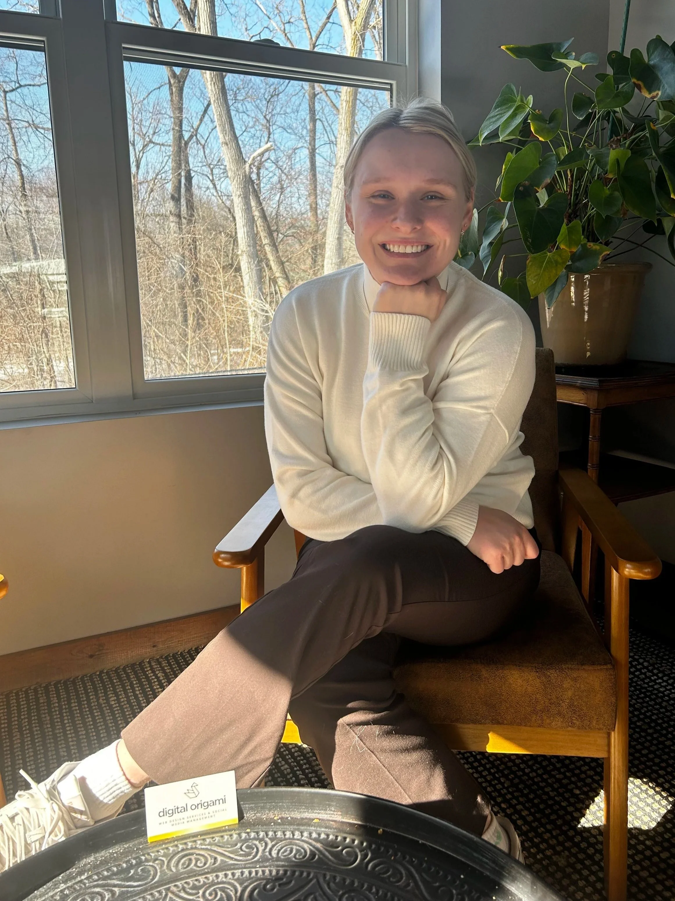 A young woman with blonde hair sitting on a wooden chair, smiling with her chin resting on her hand, near a window with a view of leafless trees. She is wearing a white sweater and brown pants, with a large potted plant on a small table nearby.