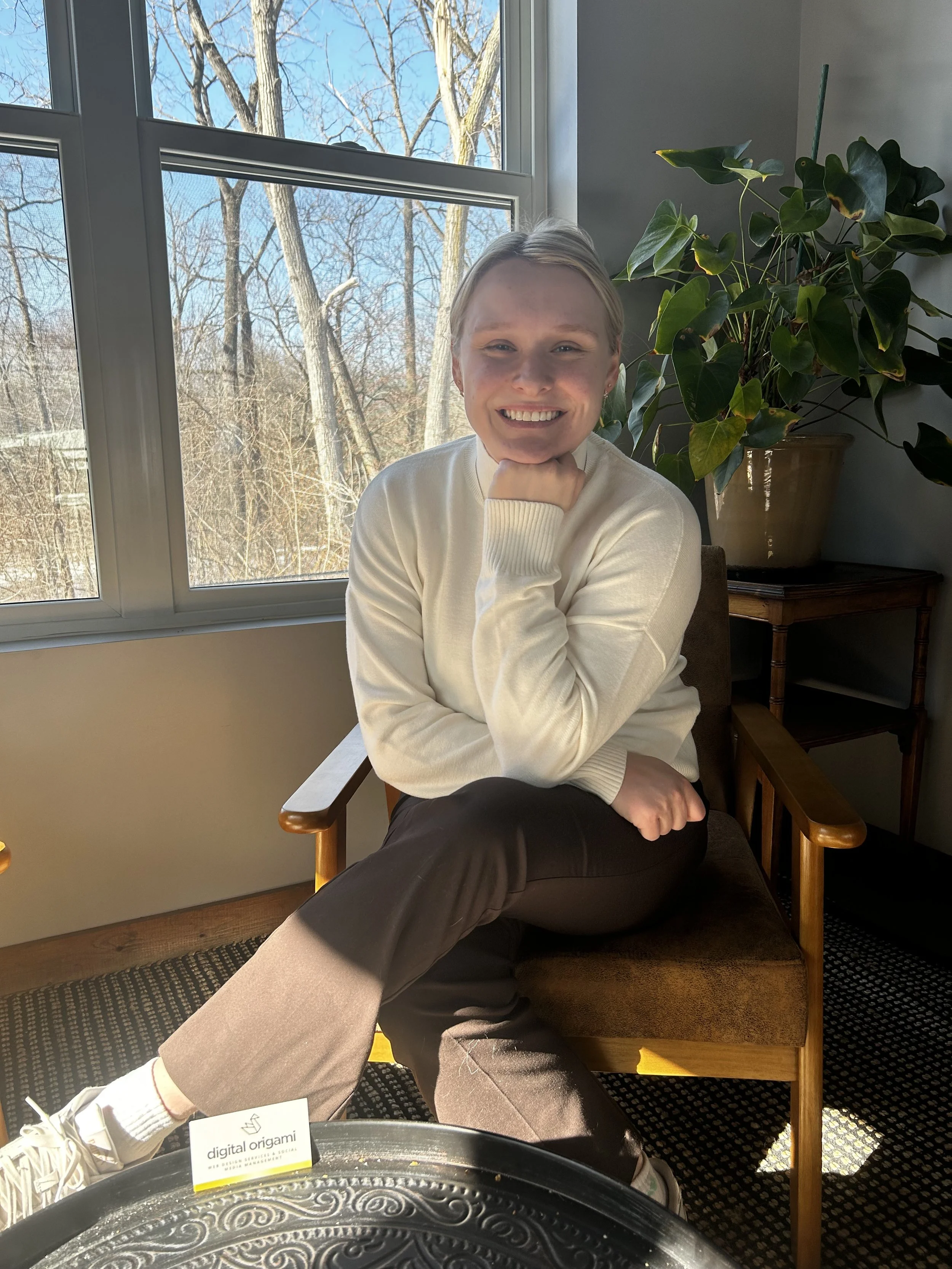 A young woman with blonde hair sitting on a wooden chair, smiling with her chin resting on her hand, near a window with a view of leafless trees. She is wearing a white sweater and dark pants, with a large potted plant on a small table nearby.