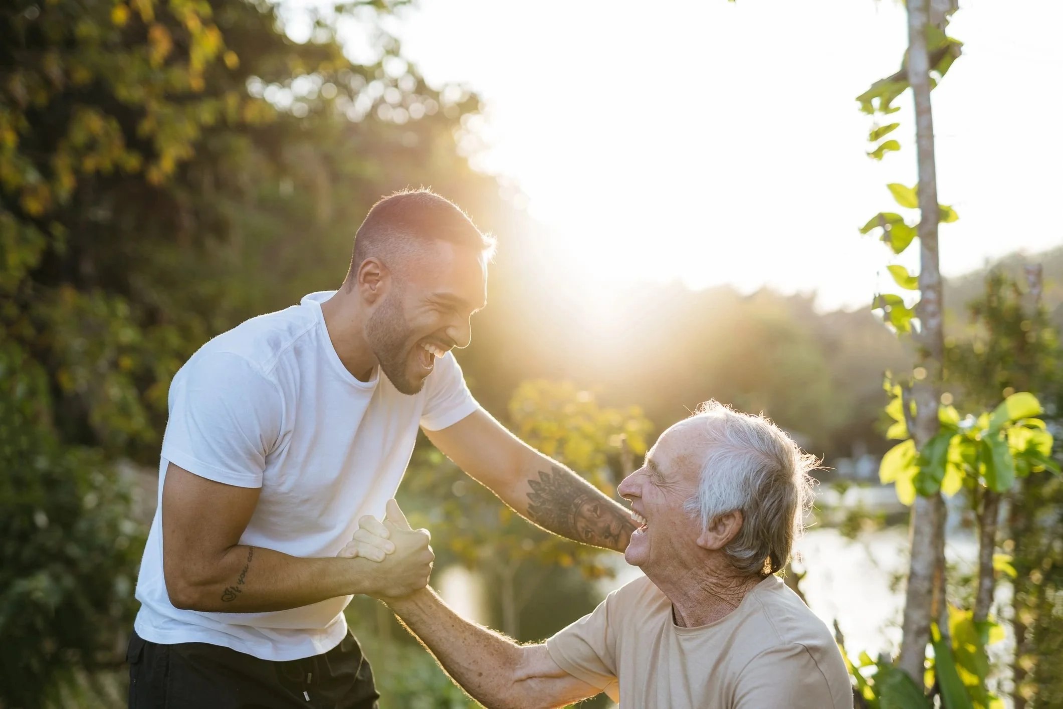 A young man and an elderly man laughing and holding hands outdoors during sunset, surrounded by green trees and plants.