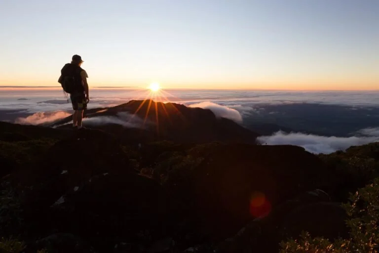 Hiker standing on a mountain at sunrise above clouds, with the sun rising over the horizon.