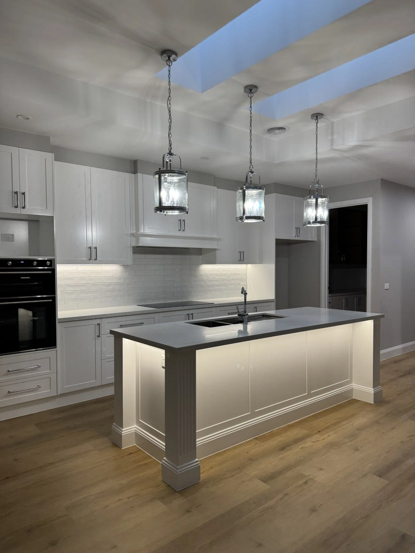 Modern kitchen with white cabinets, a kitchen island with a lighted base, three hanging pendant lights, a white subway tile backsplash, and natural wood flooring.