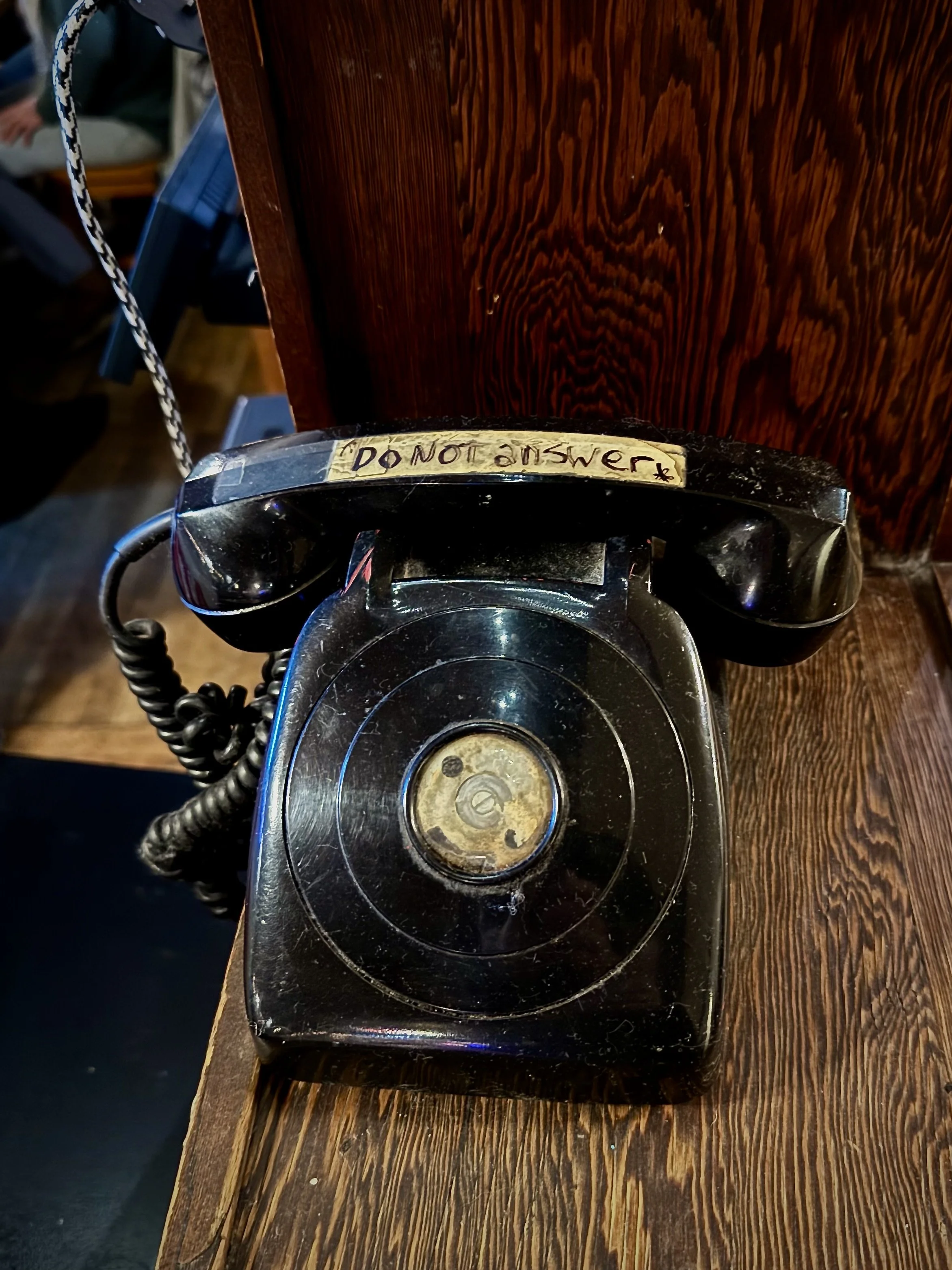 Old black rotary phone with a worn dial, placed on a wooden surface, with a handwritten note saying 'Do Not Answer' taped above the receiver.