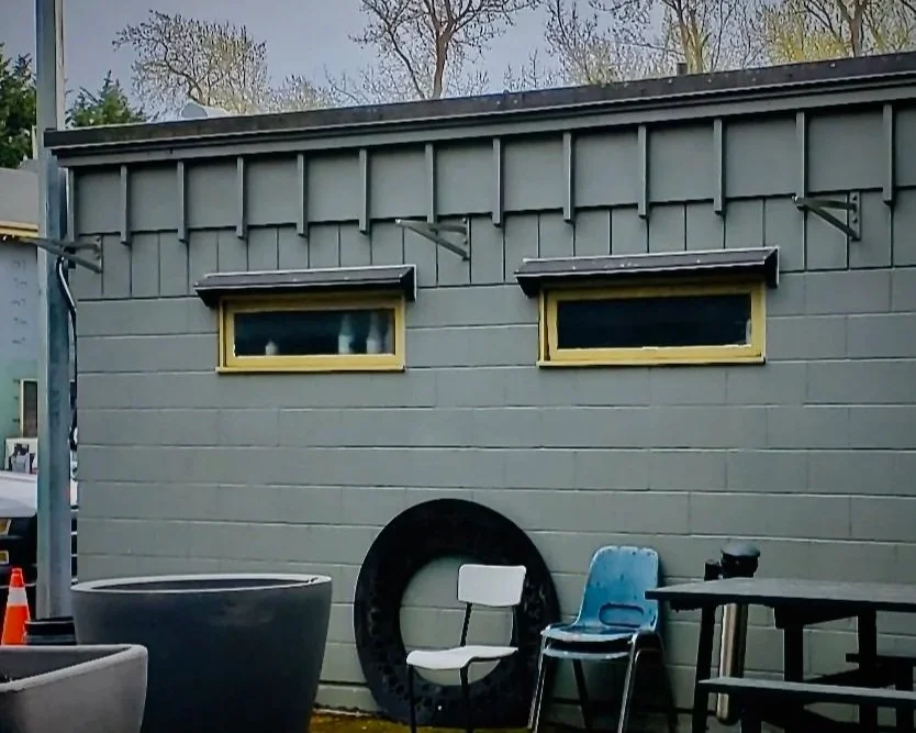 Exterior of a gray building with two small rectangular windows with yellow frames, surrounded by outdoor chairs and a large rubber tire leaning against the wall.