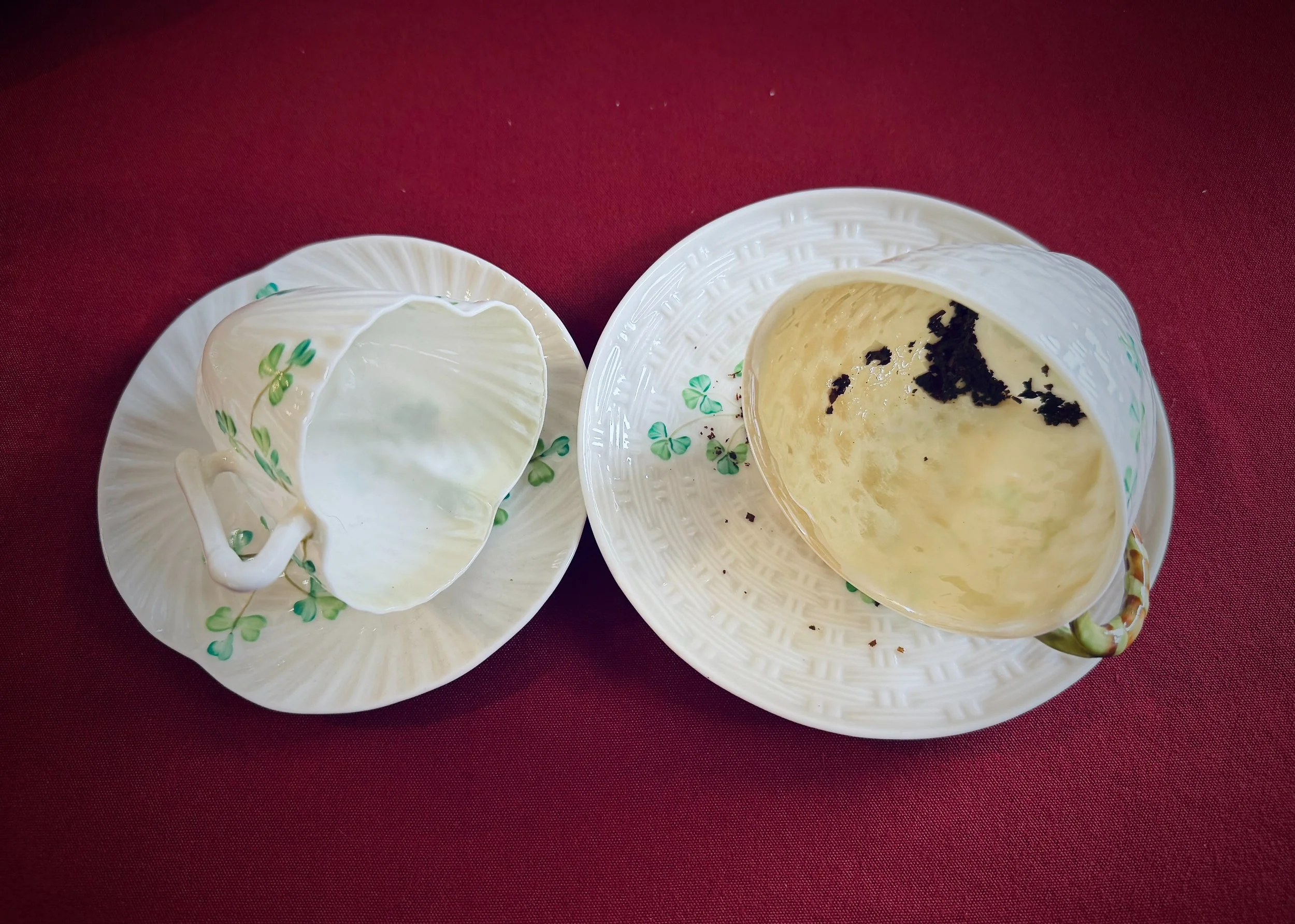 Two cups with green shamrock designs on decorative saucers sit on a dark red tablecloth. One cup is empty, while the other has remnants of tea leaves from a reading.