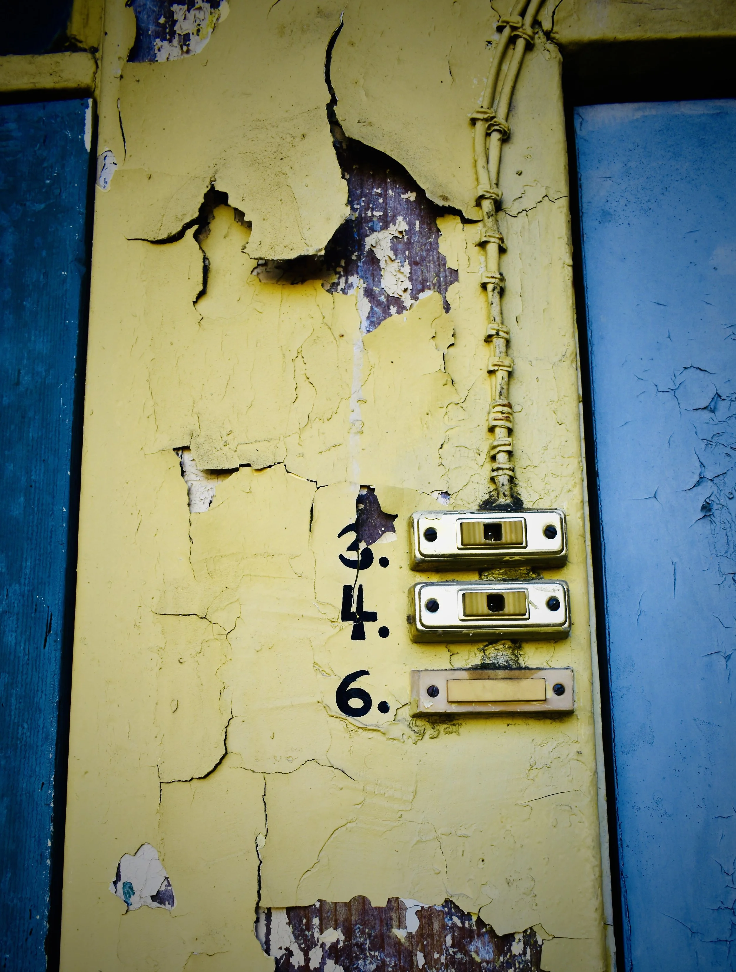Close-up of an old yellow wall with peeling paint, cracked after exposure to weather. Two vintage, beige-coloured light switches are mounted on the wall, along with a yellow electrical conduit running vertically.  The numbers 3, 4 and 6 are visible.