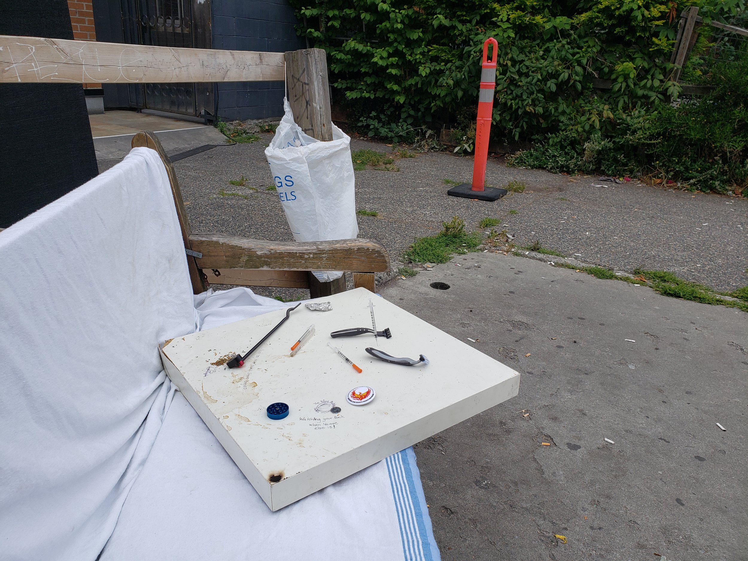Outdoor scene with a white mattress, a white board on top with tools and small items, a white trash bag, a wooden bench, and an orange traffic cone on a sidewalk next to green bushes and trees.