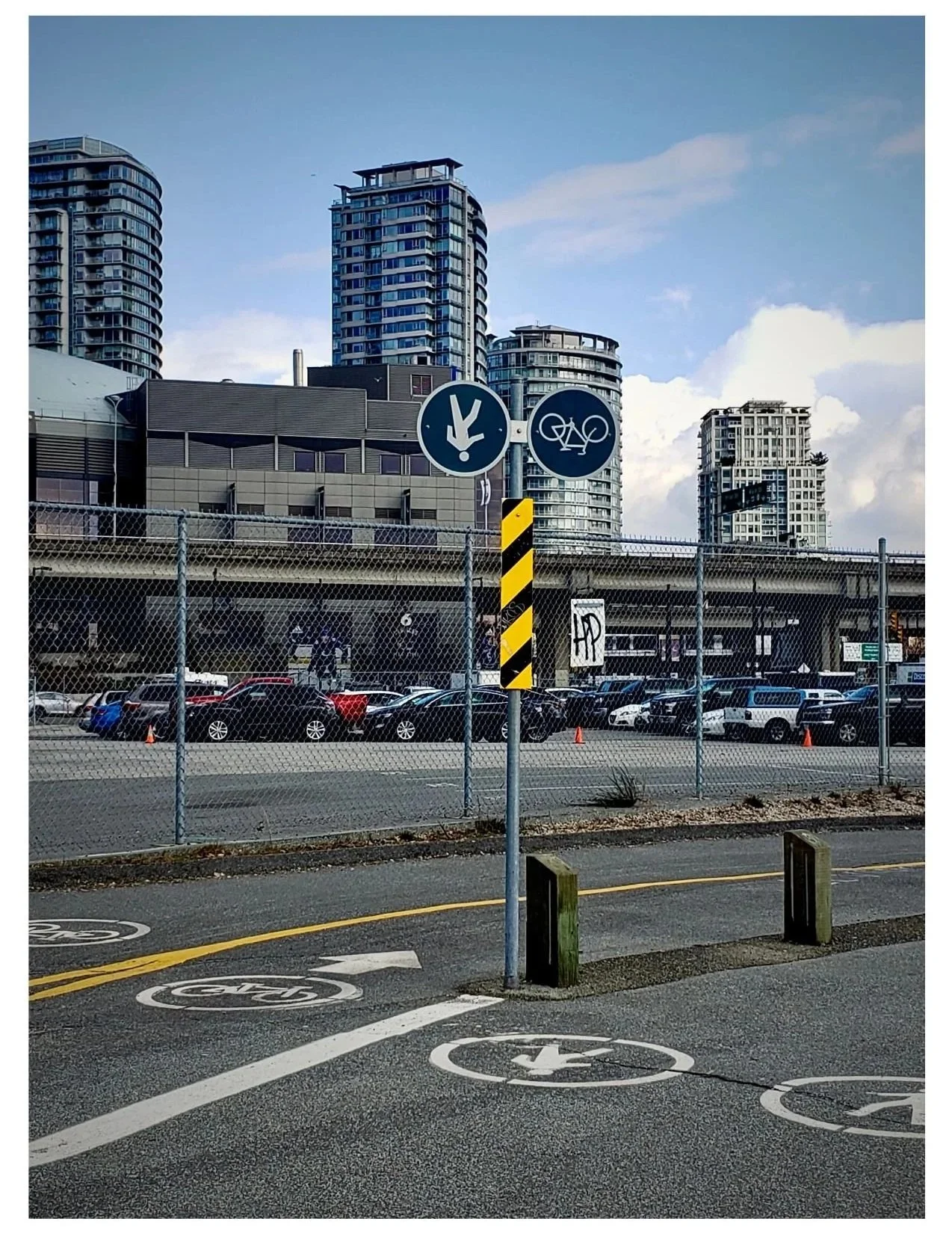 Urban parking area and bike lane with city buildings in the background. Fence separates parking lot with cars from bike lane marked on street, with an upside down sign indicating bike lane and direction.