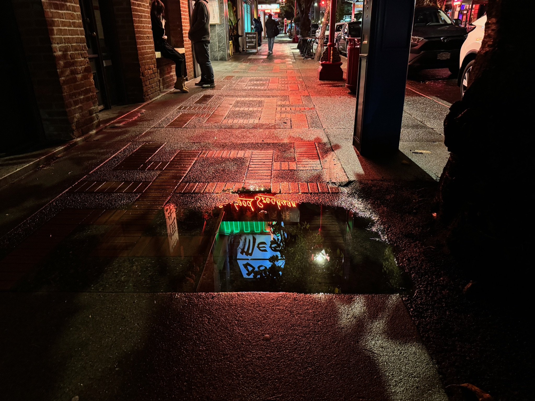 City sidewalk at night with reflections of neon signs in a puddle, brick buildings, streetlights, and parked cars. Pedestrians are visible in the distance.