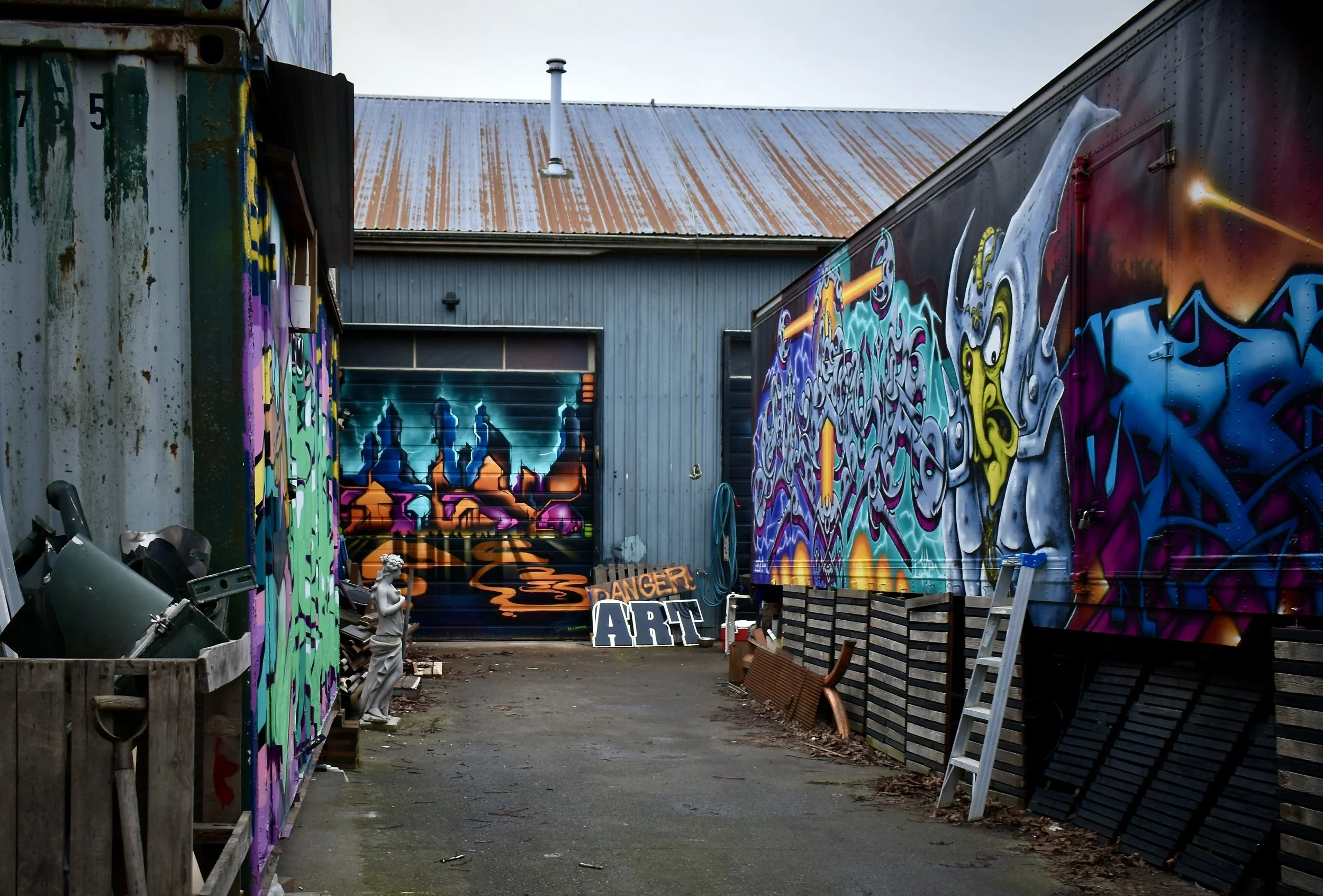 Graffiti art murals on a garage door and walls of an alleyway, with a building in the background, some trash and tools on the ground, and a statue of a person on the left side.