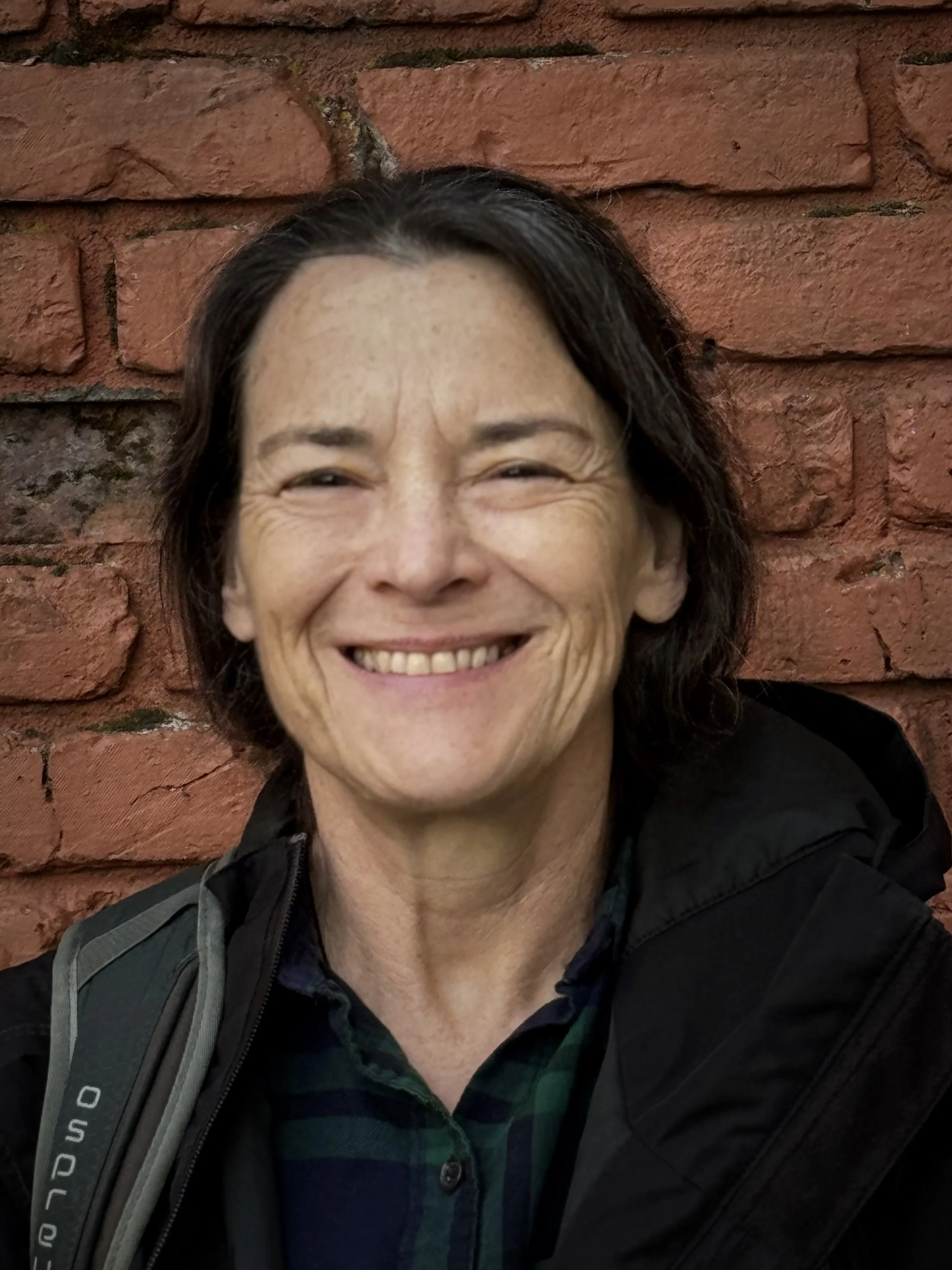 A smiling woman with dark hair and a checked shirt, standing in front of a red brick wall.
