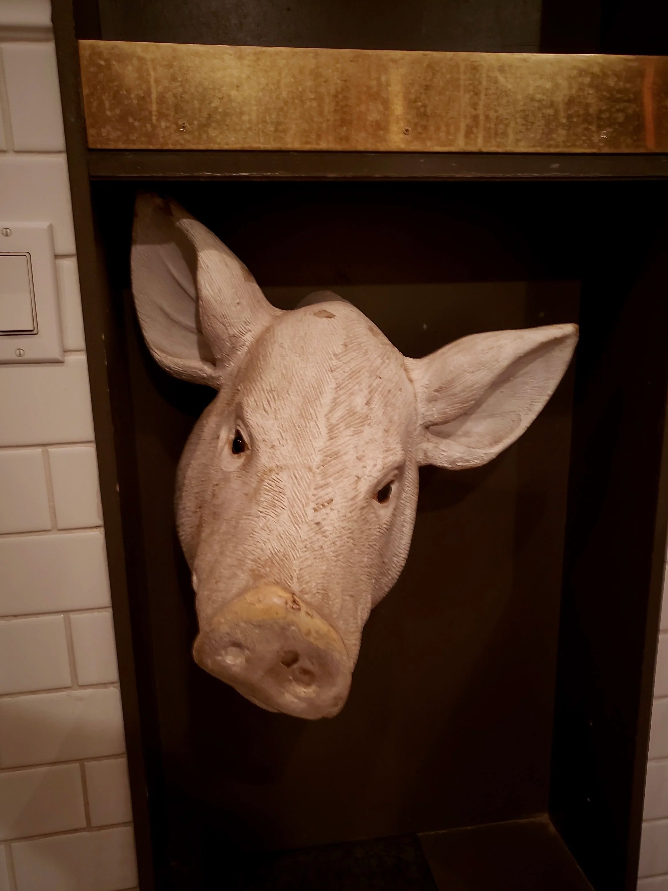 Wooden pig head sculpture mounted inside a dark display case in a room with tiled walls.