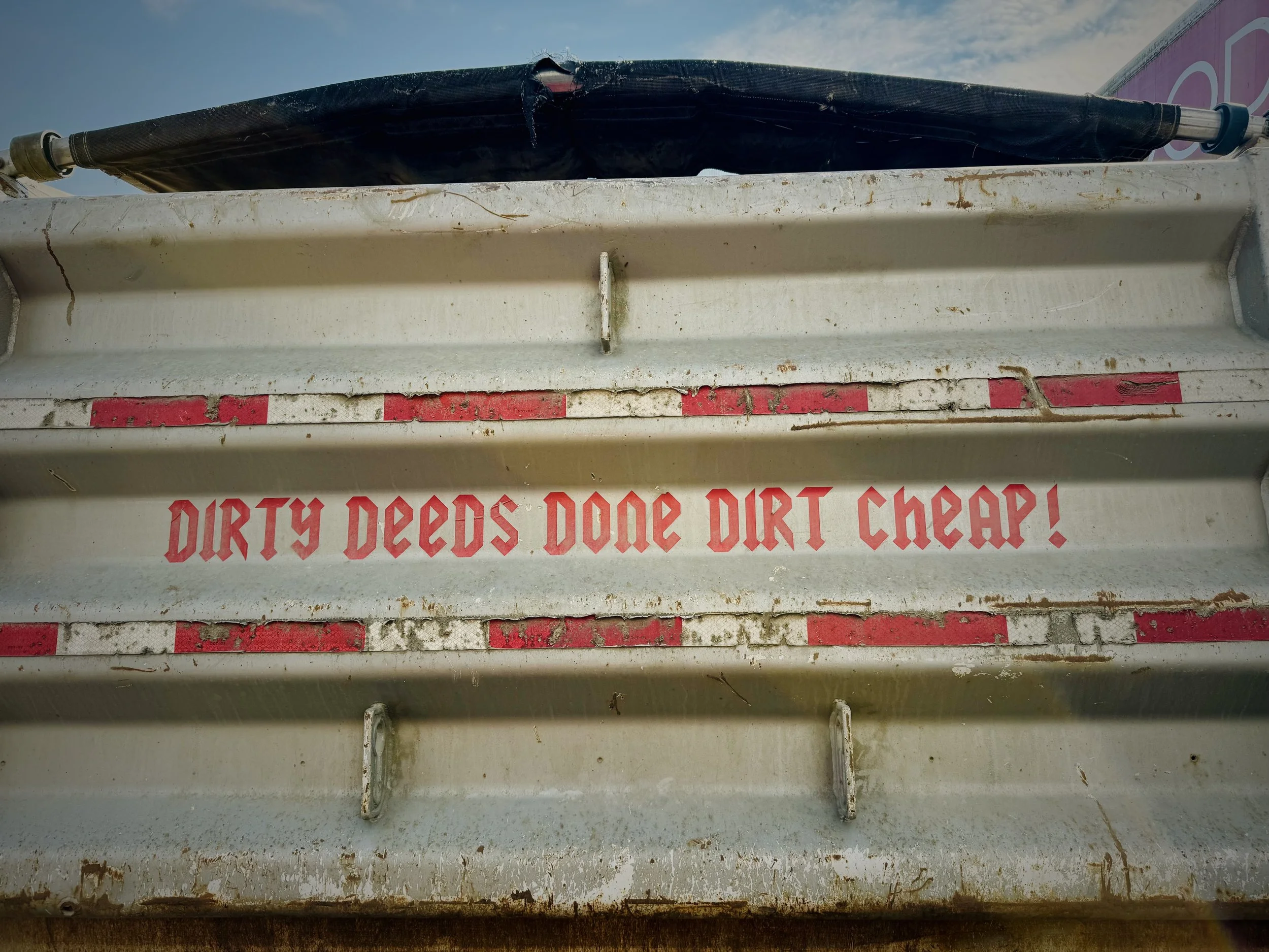Close-up of a rusty, dirty white trailer with red text that says 'DIRTY DEEDS DONE DIRT CHEAP!'