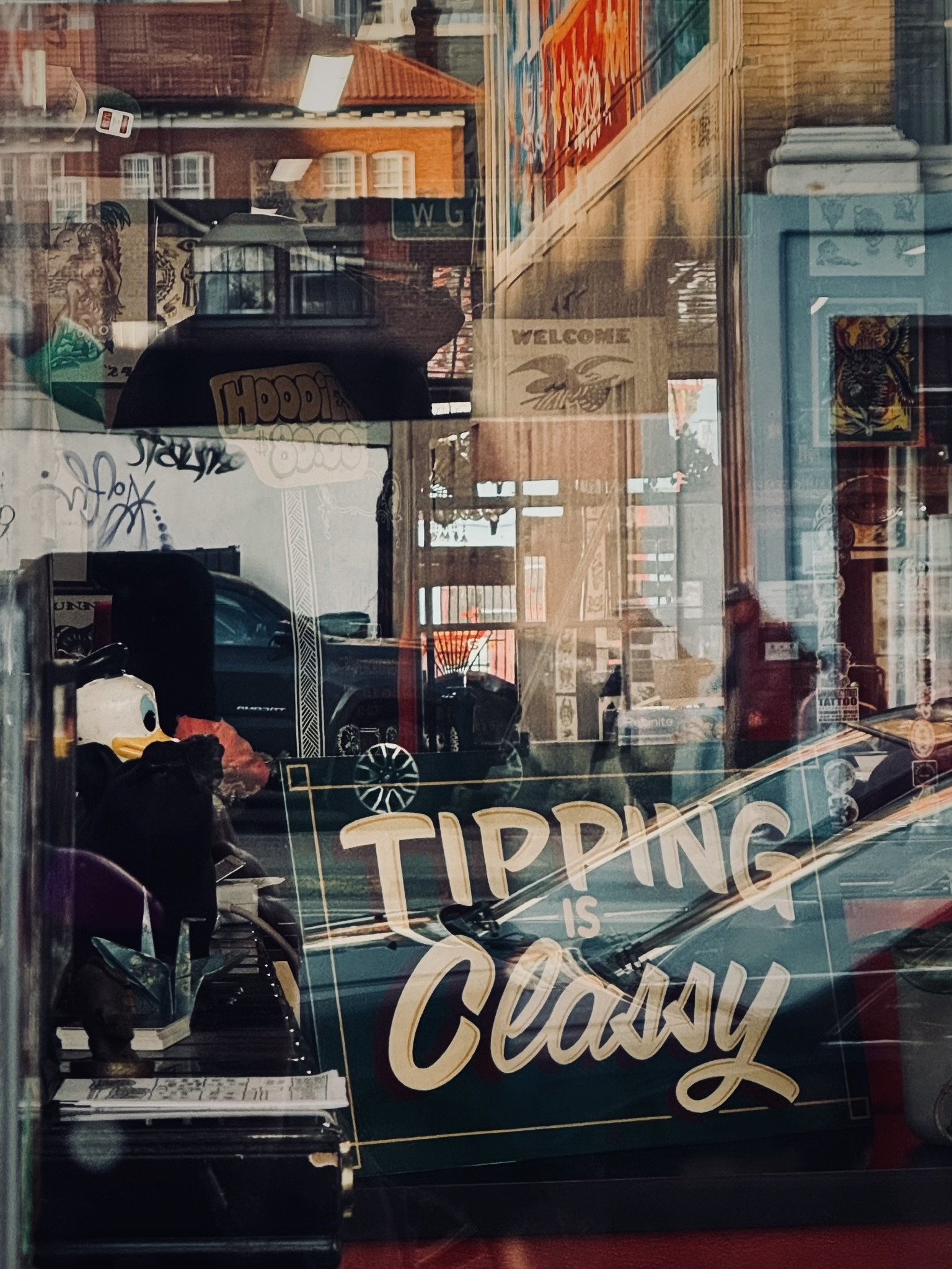 A view through a store window with a sign that reads 'TIPPING IS CLASSY'. Reflections show parked cars, buildings, and signs outside, with visible signage including a welcome sign and graffiti inside.