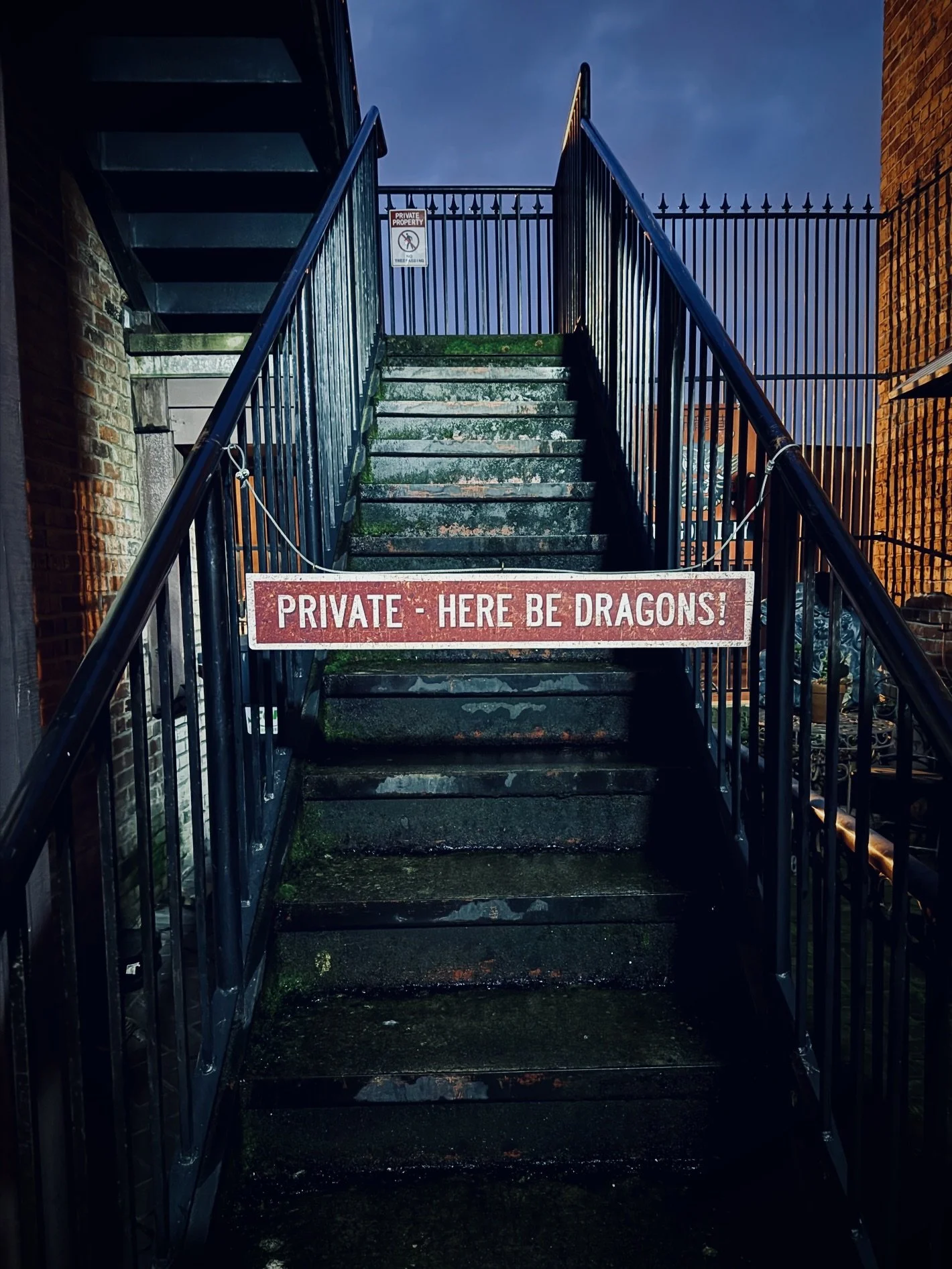 An outdoor metal staircase with a sign that reads 'Private - Here Be Dragons!' blocking the entrance, flanked by brick buildings on both sides during dusk.