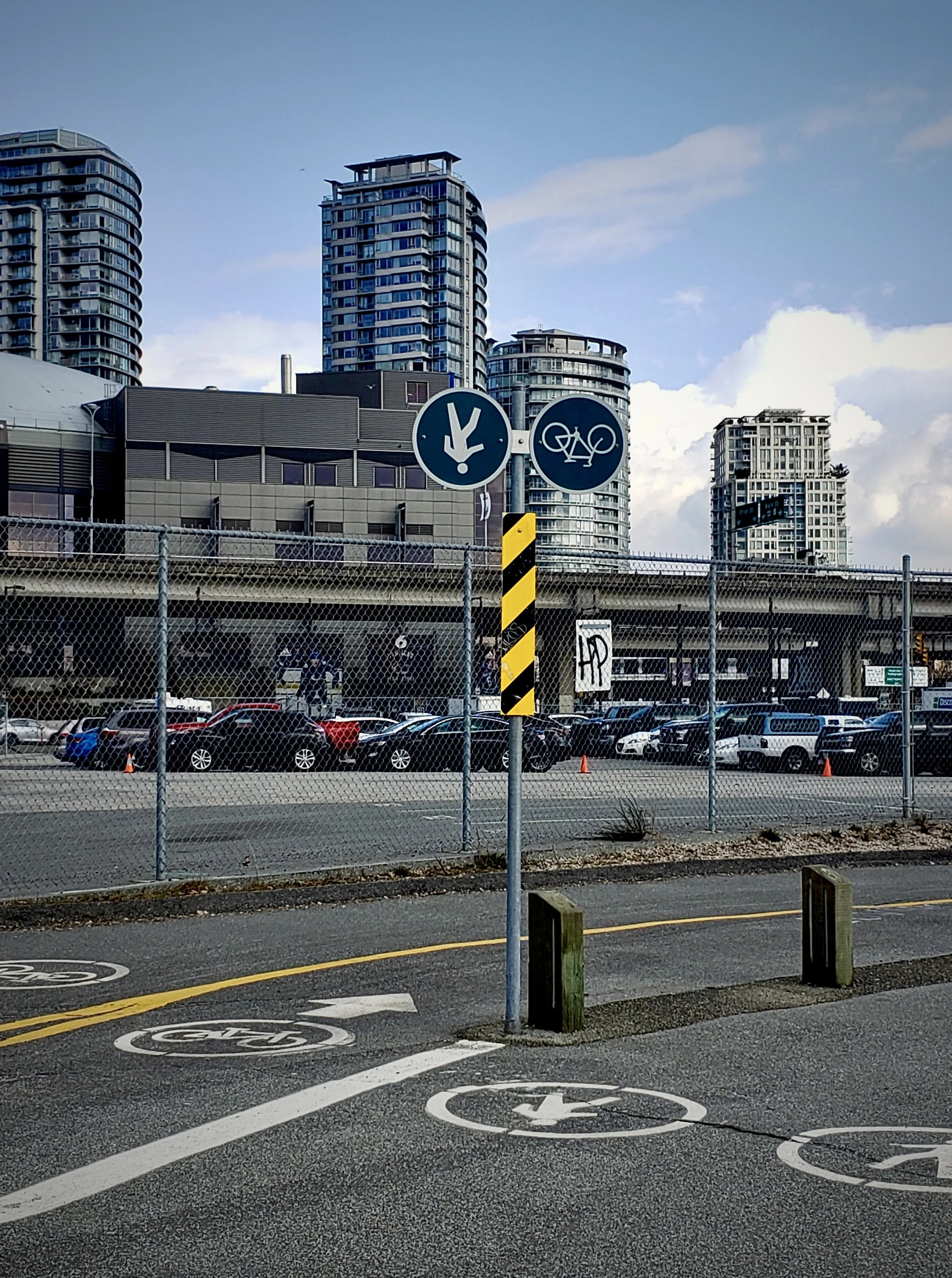 Urban street scene with parking lot, high-rise buildings, with an upside down sign for bicycles and pedestrians, a chain-link fence, and a bike lane.