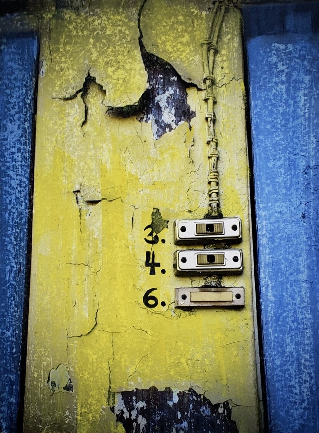 Close-up of an old, peeling yellow wall with cracked paint, rusted electrical wires, and broken switches, framed by blue trim.